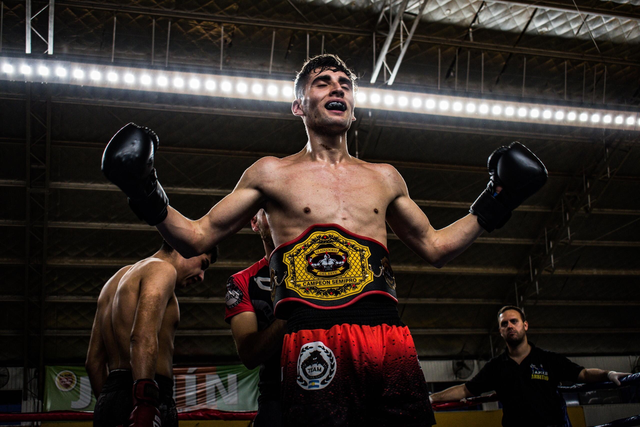 Boxeador con el cinturón de campeón y los guantes en alto en señal de victoria, de pie en el ring con sus rivales y el árbitro al fondo.