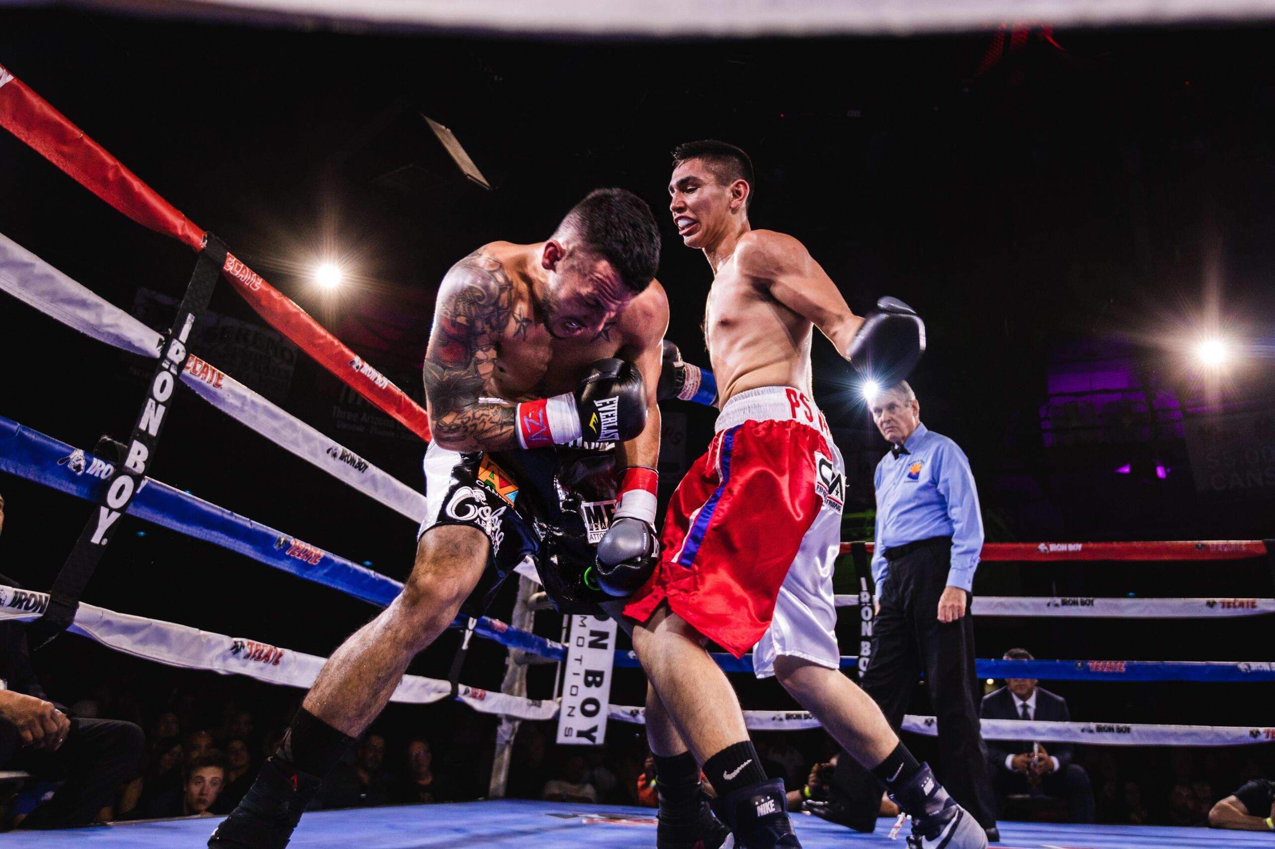 Dos boxeadores en un ring durante un combate; uno de ellos asesta un puñetazo al abdomen del otro, mientras el árbitro observa atentamente.