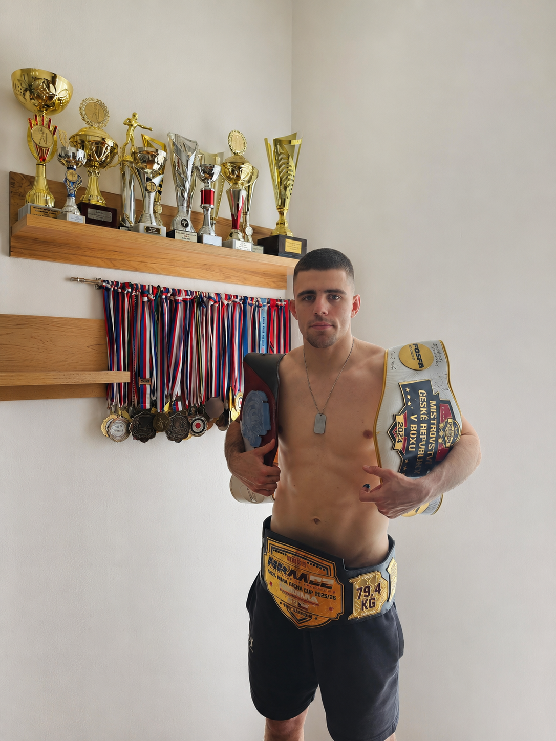 Un deportista sin camiseta posa con aire seguro junto a tres cinturones de campeón, rodeado de numerosos trofeos y medallas colocados en las estanterías de la pared.