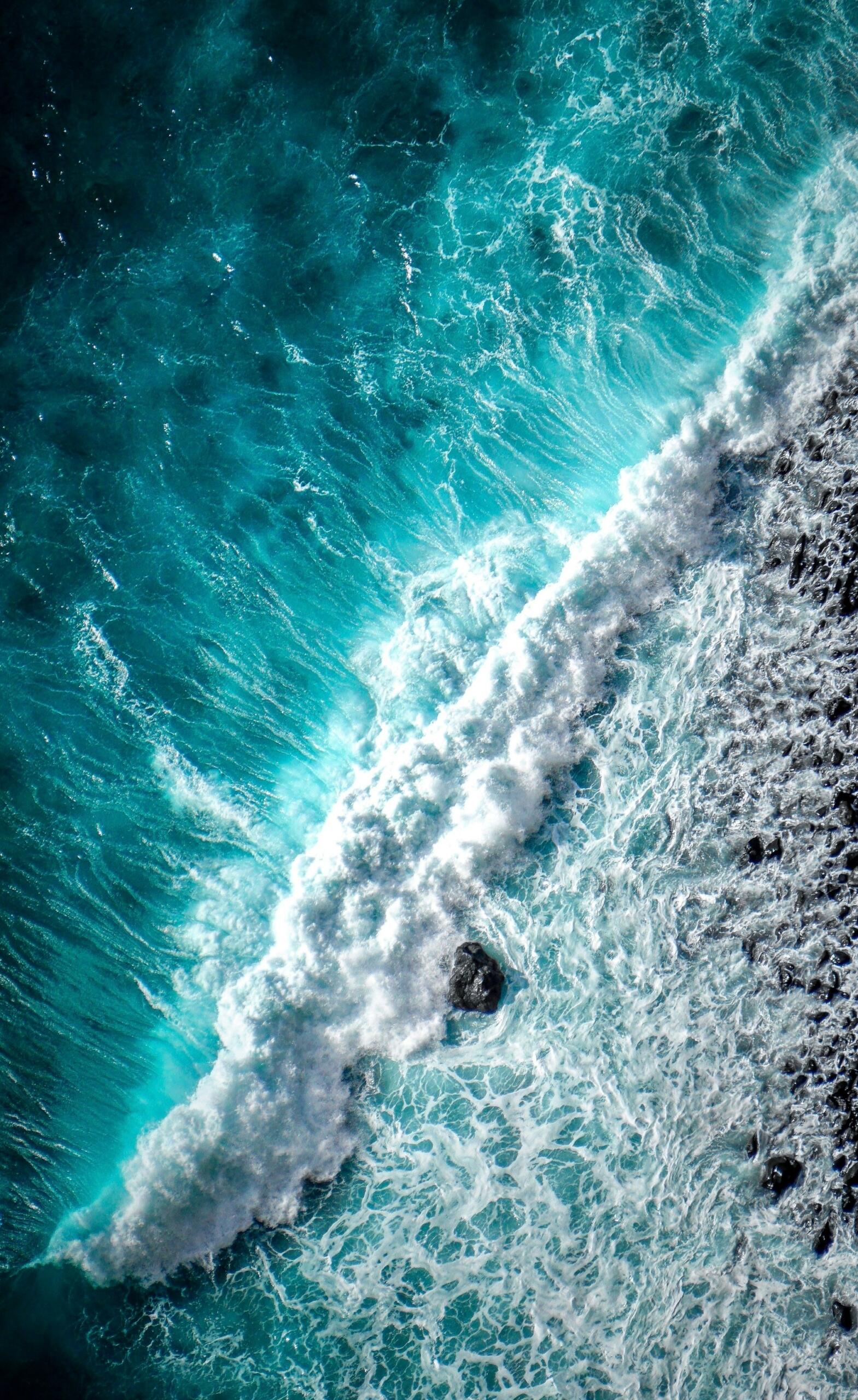 Vista aérea de las olas turquesas del océano rompiendo contra una costa rocosa, creando una espuma blanca que se arremolina y una escena costera dinámica y poderosa.