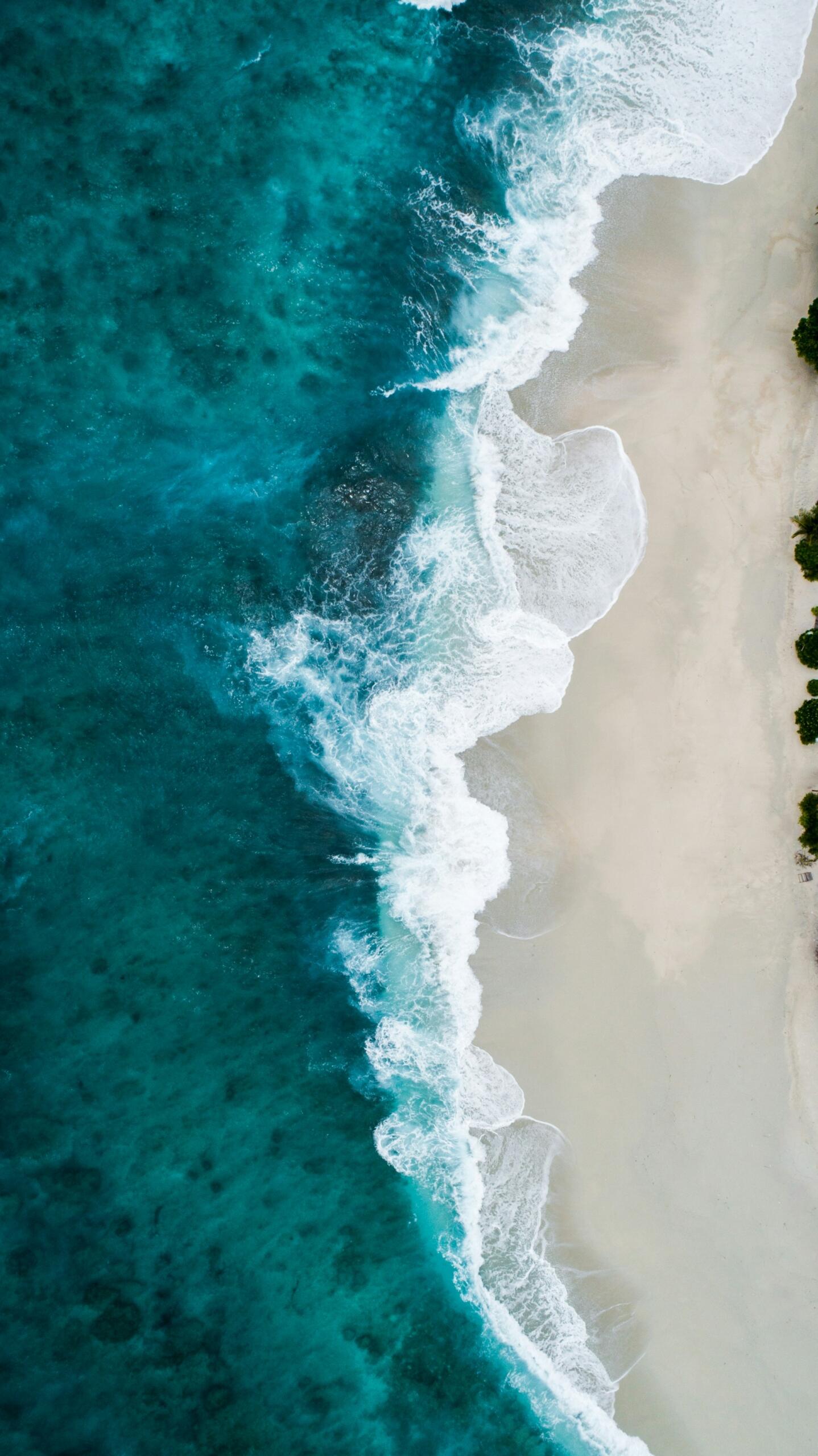Vista aérea de las olas turquesas del mar rompiendo en una playa de arena clara, creando motivos blancos y espumosos en un sereno paisaje costero tropical.