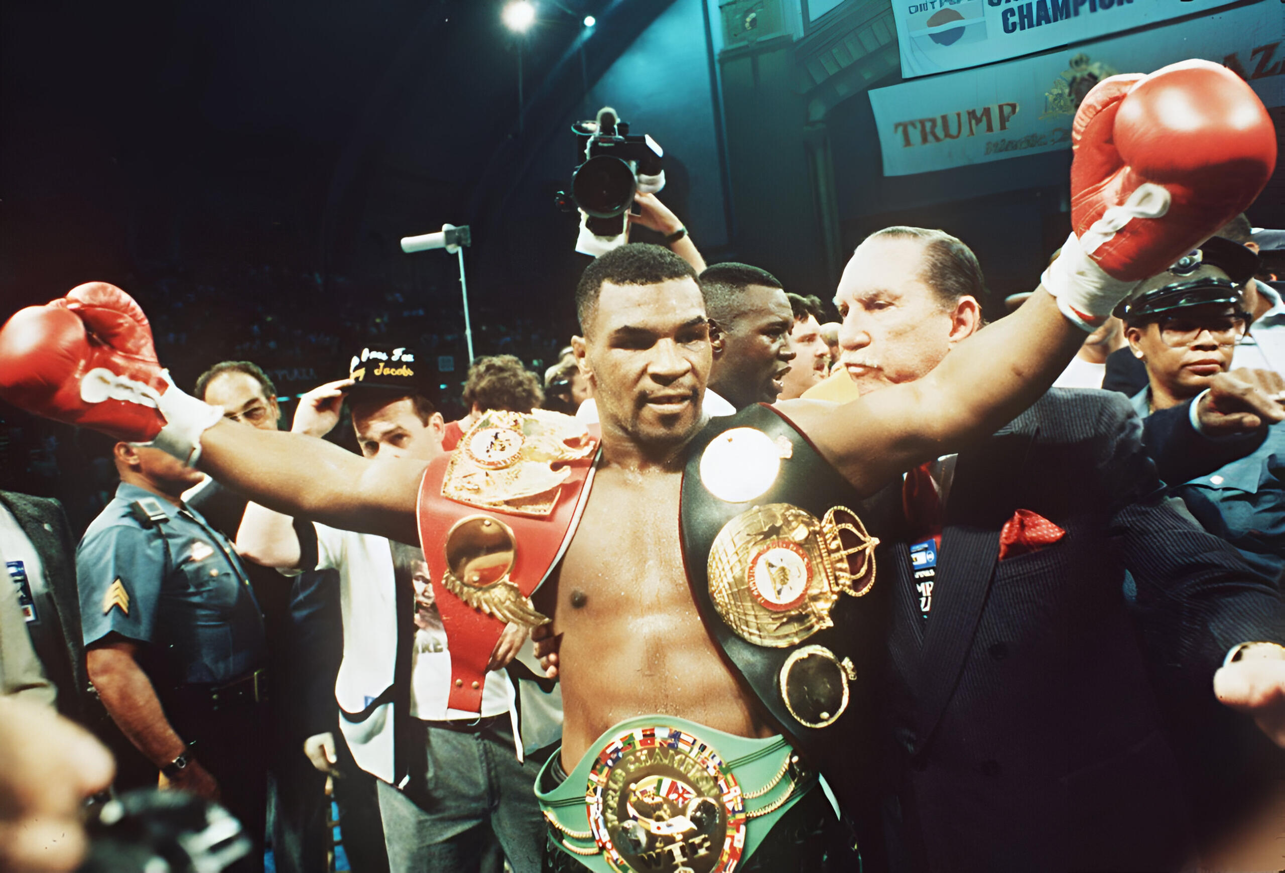 El boxeador Mike Tyson celebrando con tres cinturones de campeón y guantes rojos en un pabellón abarrotado tras un combate.