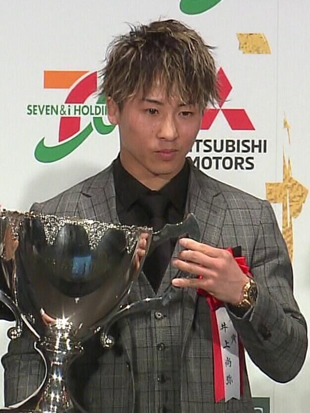 Joven con un traje gris a cuadros que sostiene un gran trofeo de plata durante una ceremonia de entrega de premios, con los logotipos de los patrocinadores al fondo.