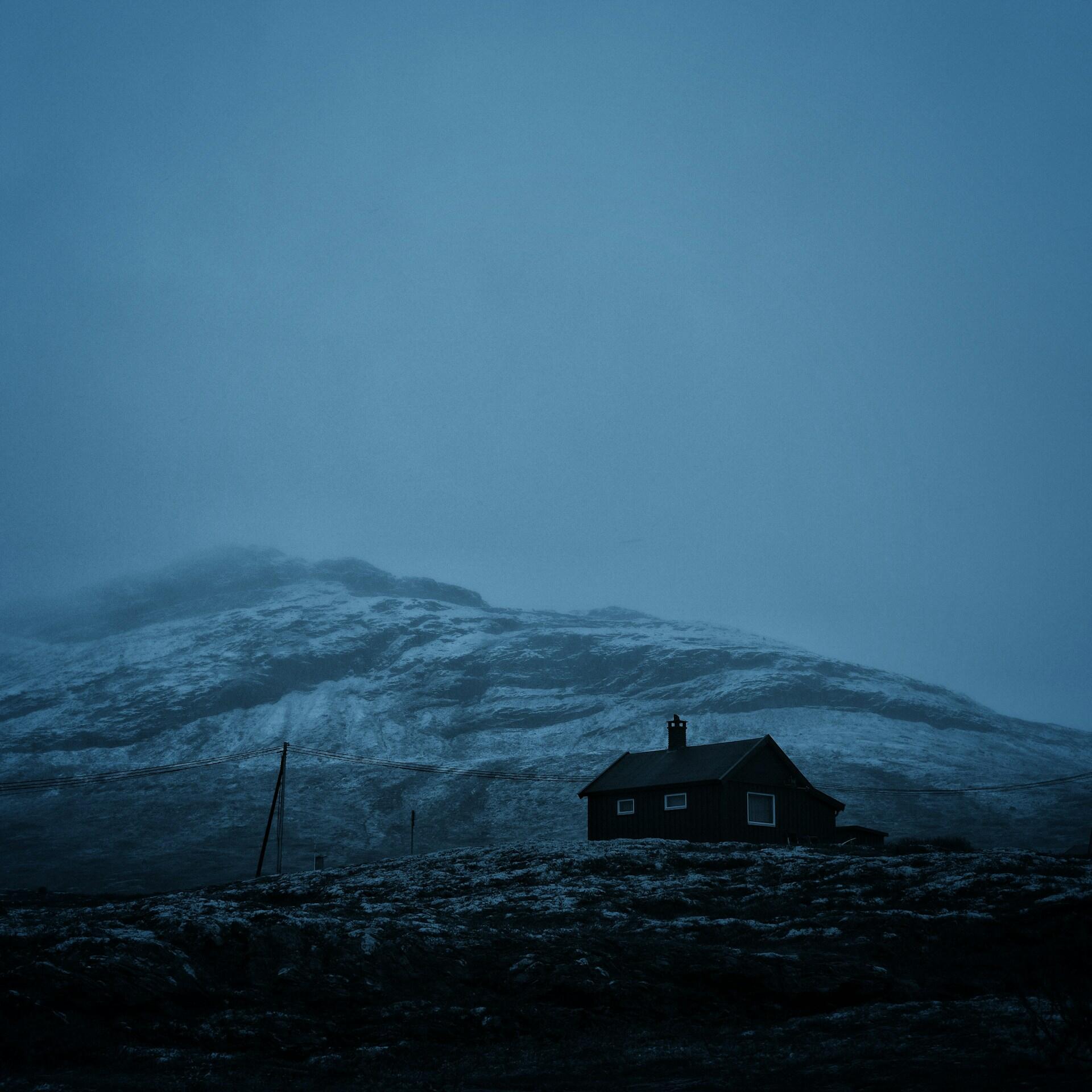 Una cabaña solitaria y oscura se alza al pie de una montaña cubierta de nieve, en el crepúsculo azul, creando una escena invernal tranquila, aislada y evocadora.
