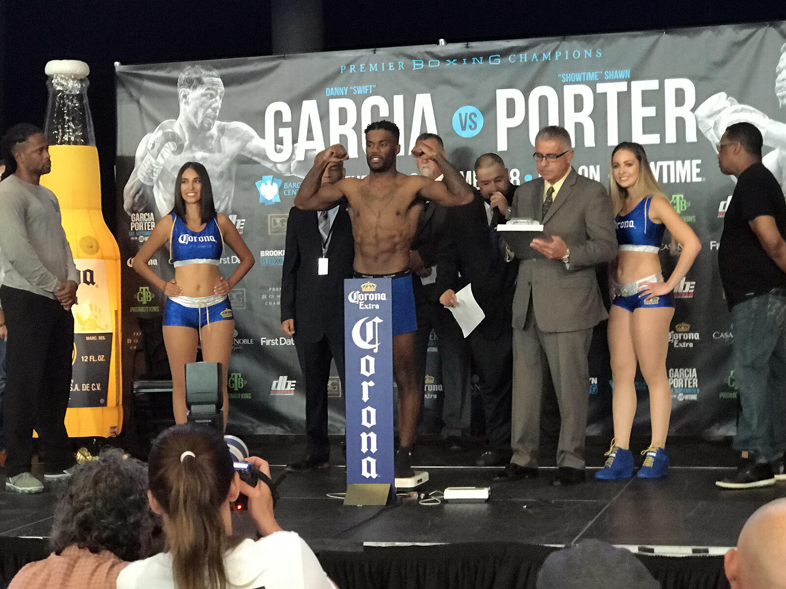 El boxeador muestra sus músculos durante el pesaje previo al combate entre García y Porter, flanqueado por las chicas del ring, vestidas con trajes azules de Corona, y por los árbitros en el escenario.