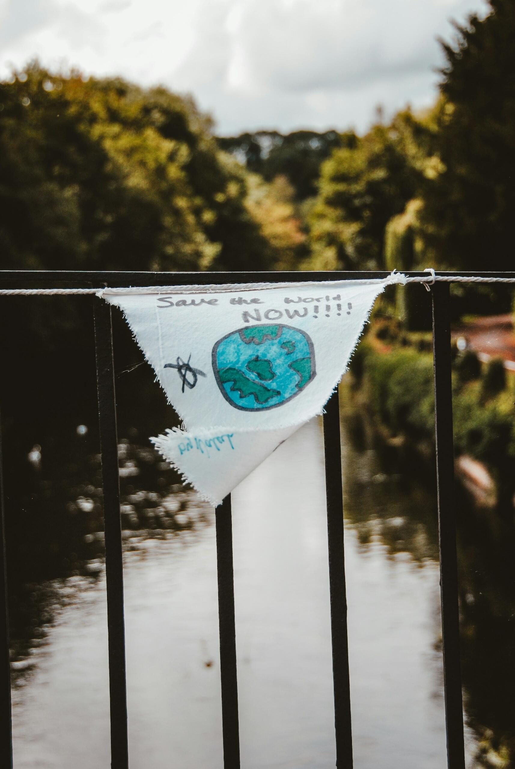 Un cartel dibujado a mano con el lema «¡¡¡Salvad el mundo YA!!!» y una Tierra de color azul verdoso cuelga de la barandilla de un puente sobre un río tranquilo, lo que evoca esperanza y preocupación por el medio ambiente.