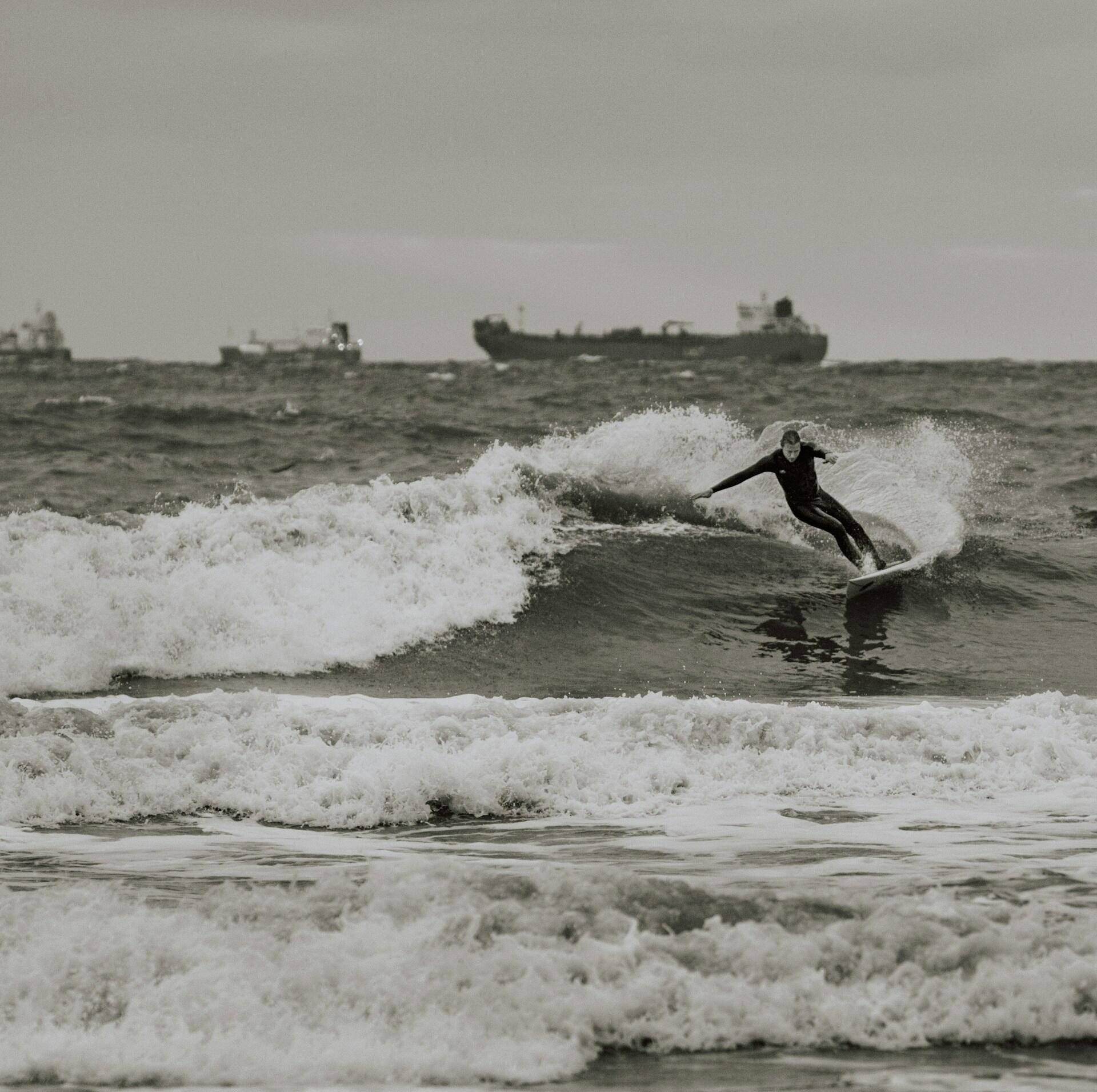 Fotografía en blanco y negro de un surfista surcando una ola que rompe, con un buque de carga en el horizonte y un mar agitado bajo un cielo gris.