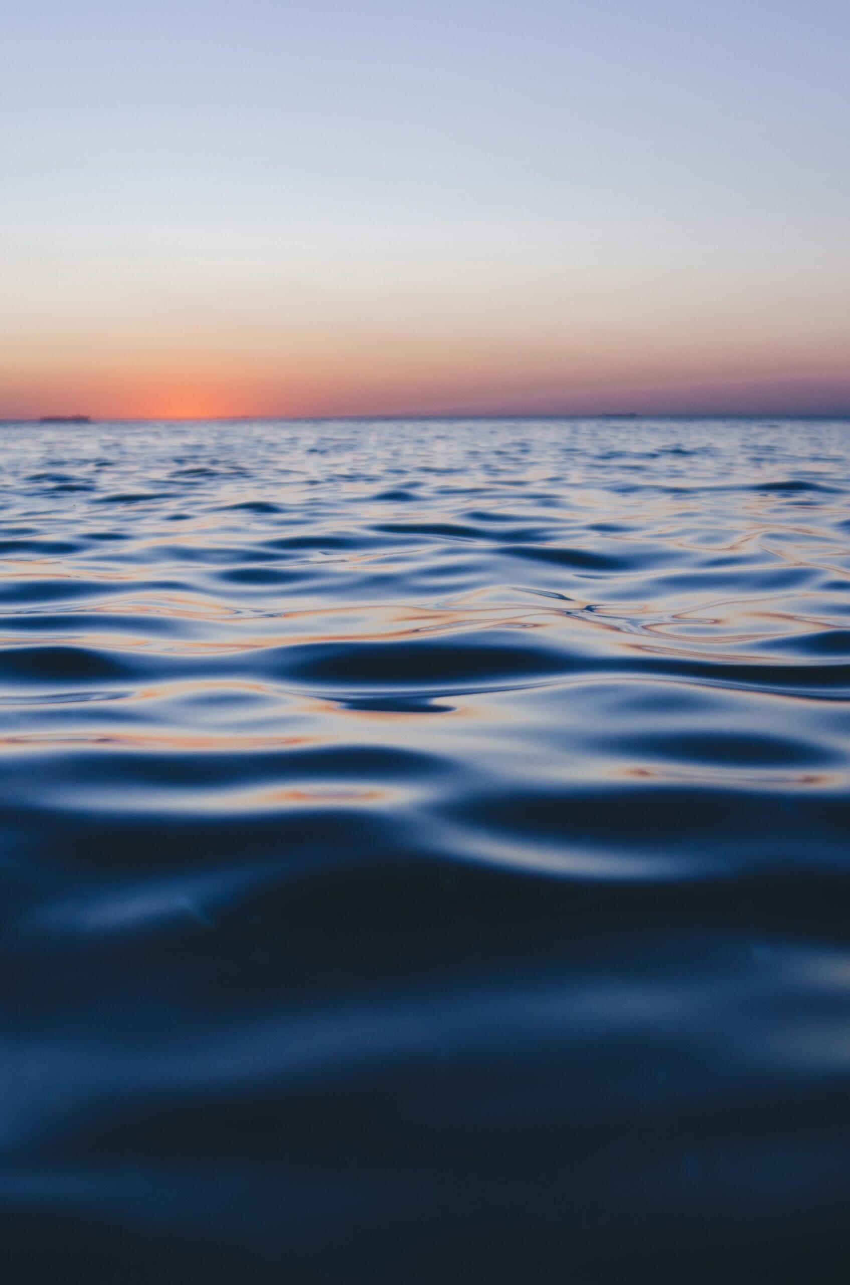 Las tranquilas olas del mar se extienden hacia el horizonte al atardecer, reflejando la luz azul y melocotón bajo un cielo en tonos pastel, creando un ambiente sereno.