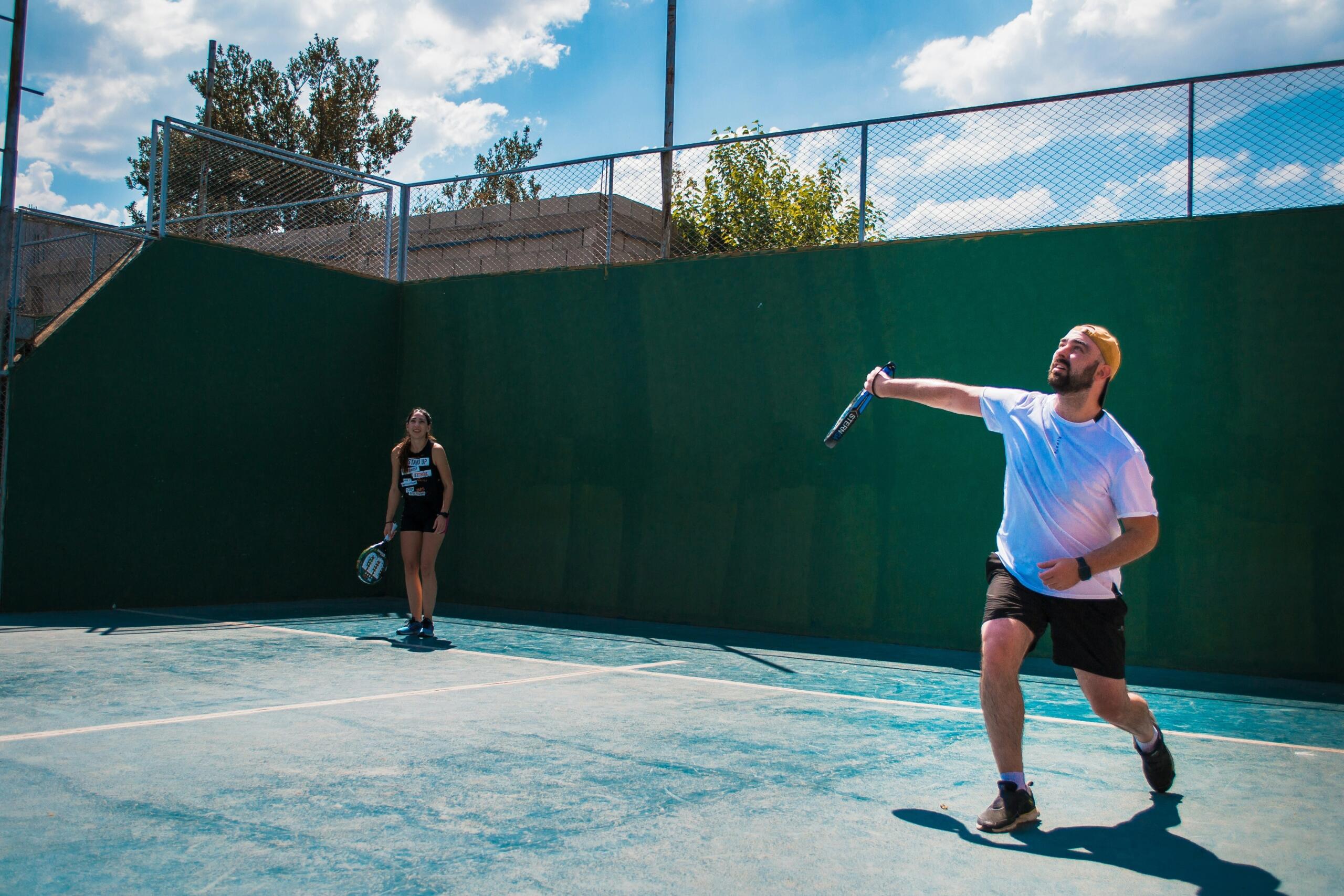 La gente juega al pickleball en una pista al aire libre rodeada por una valla metálica, bajo un cielo soleado y nublado.