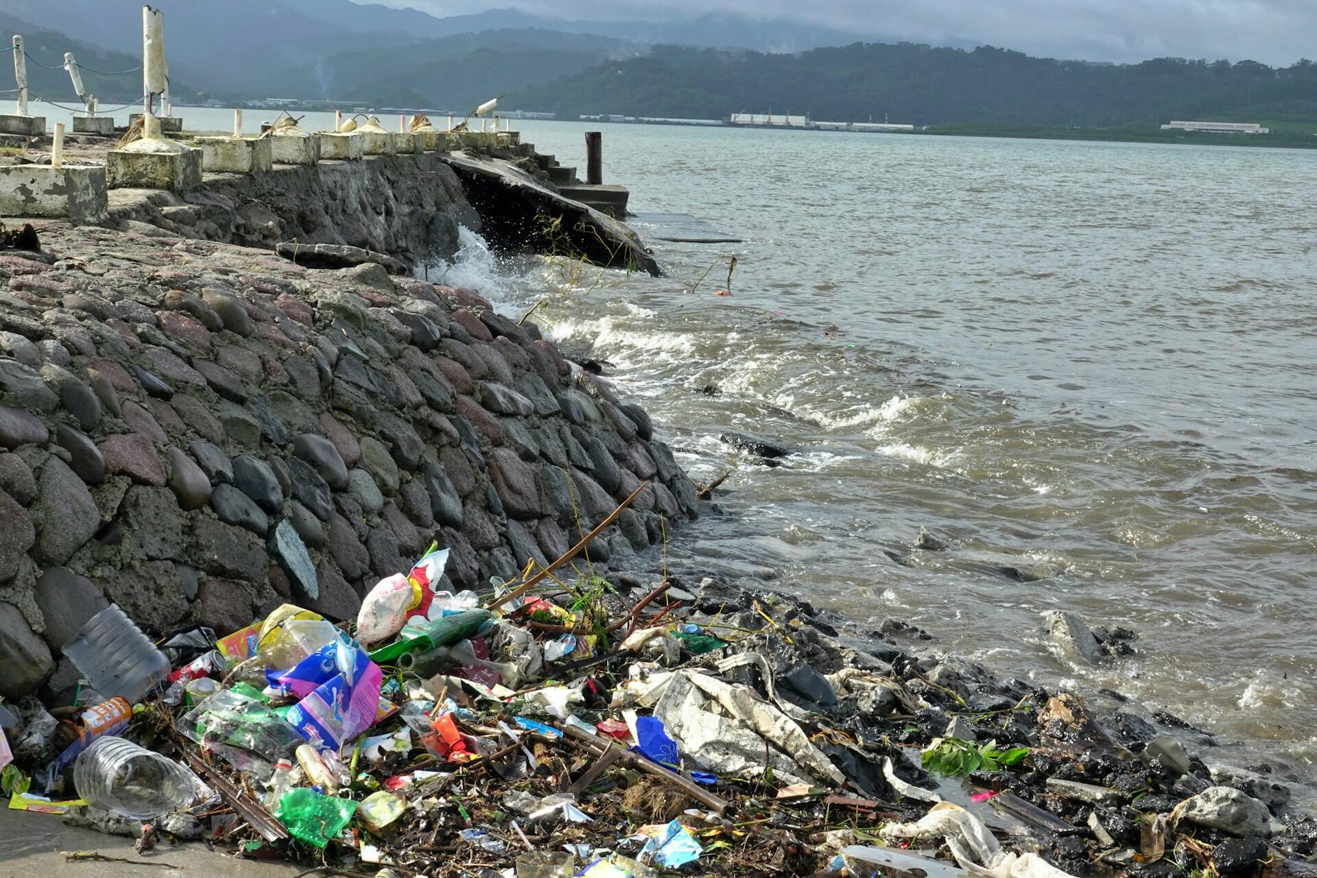 Costa rocosa llena de basura junto a aguas agitadas, con botellas de plástico y escombros en primer plano, un muelle y montañas envueltas en niebla al fondo.