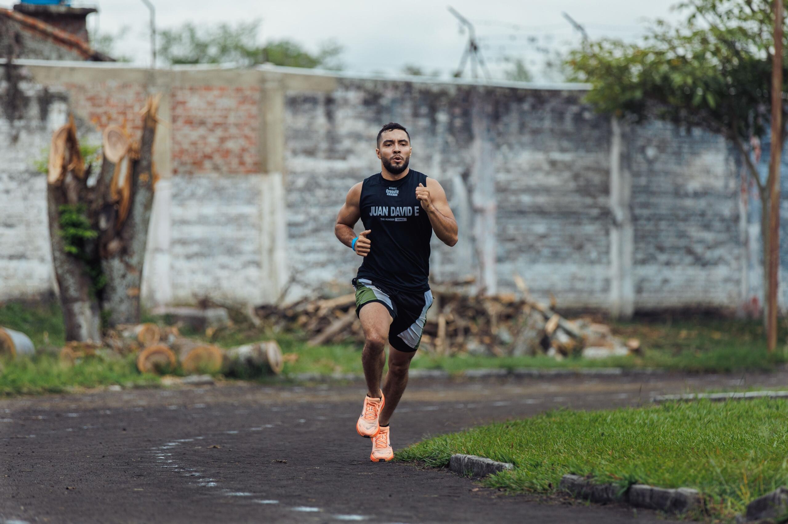 Un hombre atlético, con una camiseta negra de CrossFit, corre por un sendero mojado junto a un muro de ladrillo desgastado por el paso del tiempo.