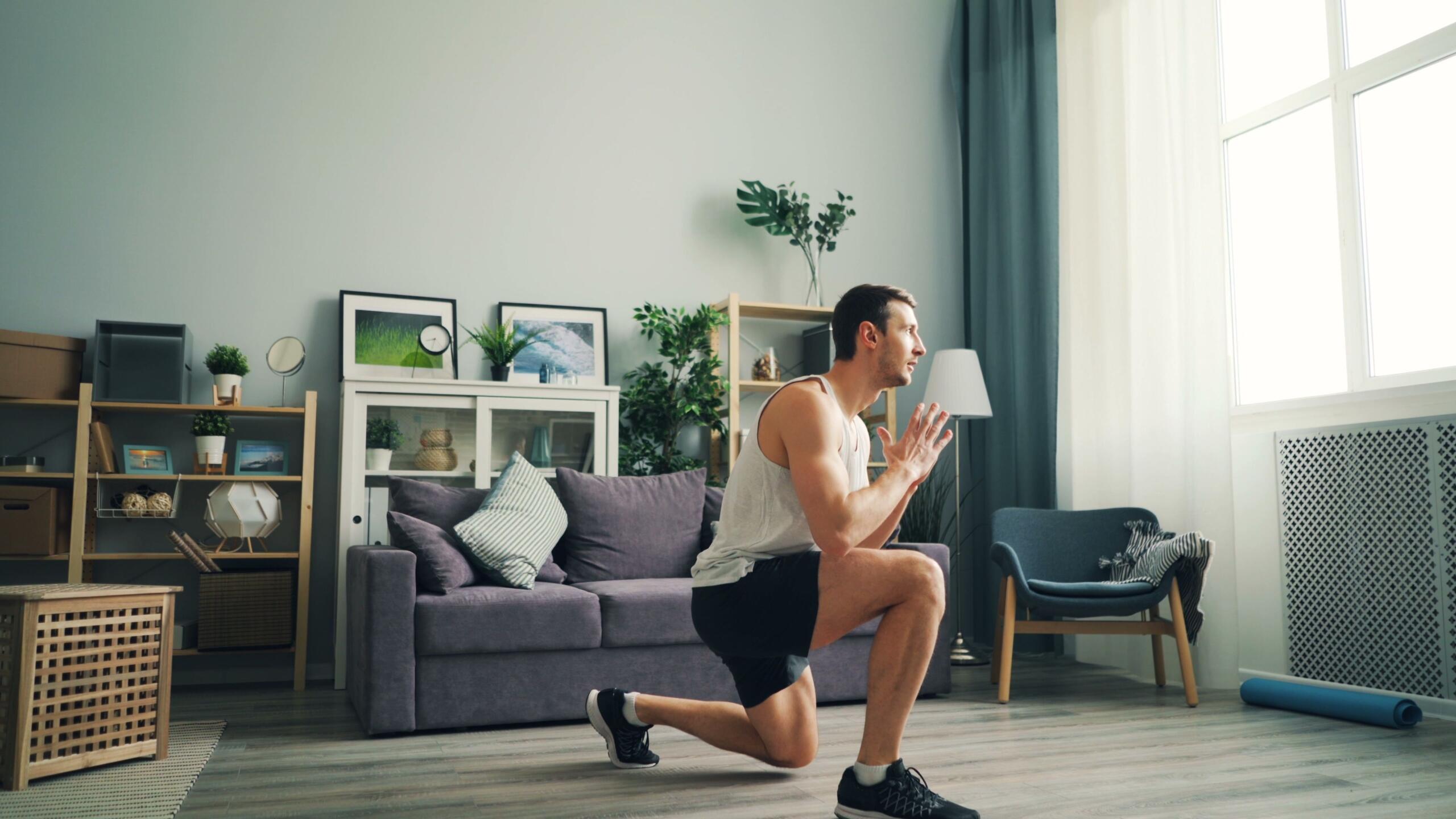 Un hombre haciendo ejercicio en el salón, realizando una zancada de rodillas junto a un sofá y unas plantas de interior.