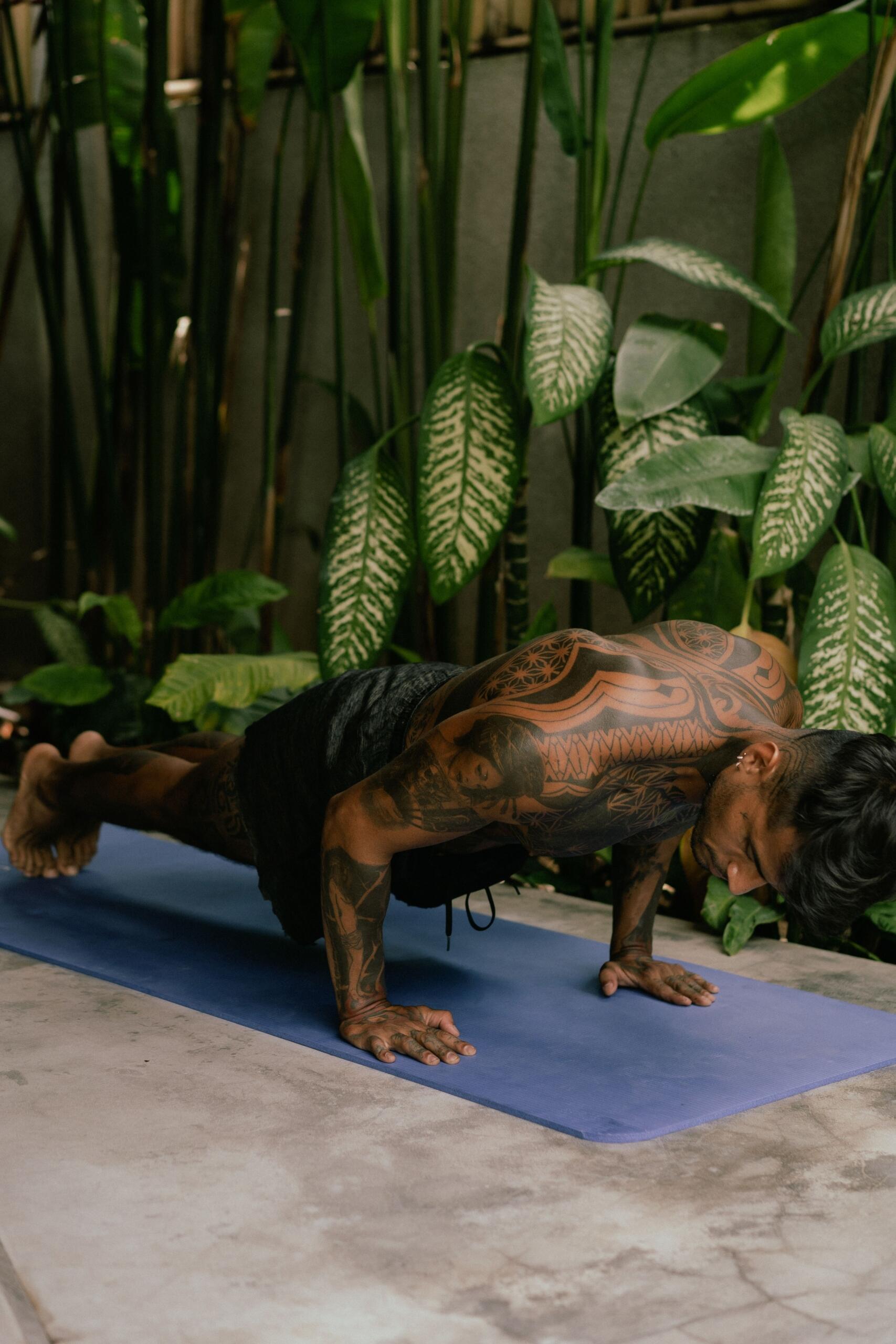 Una persona tatuada realiza una postura de yoga sobre una esterilla azul, rodeada de exuberantes plantas tropicales.