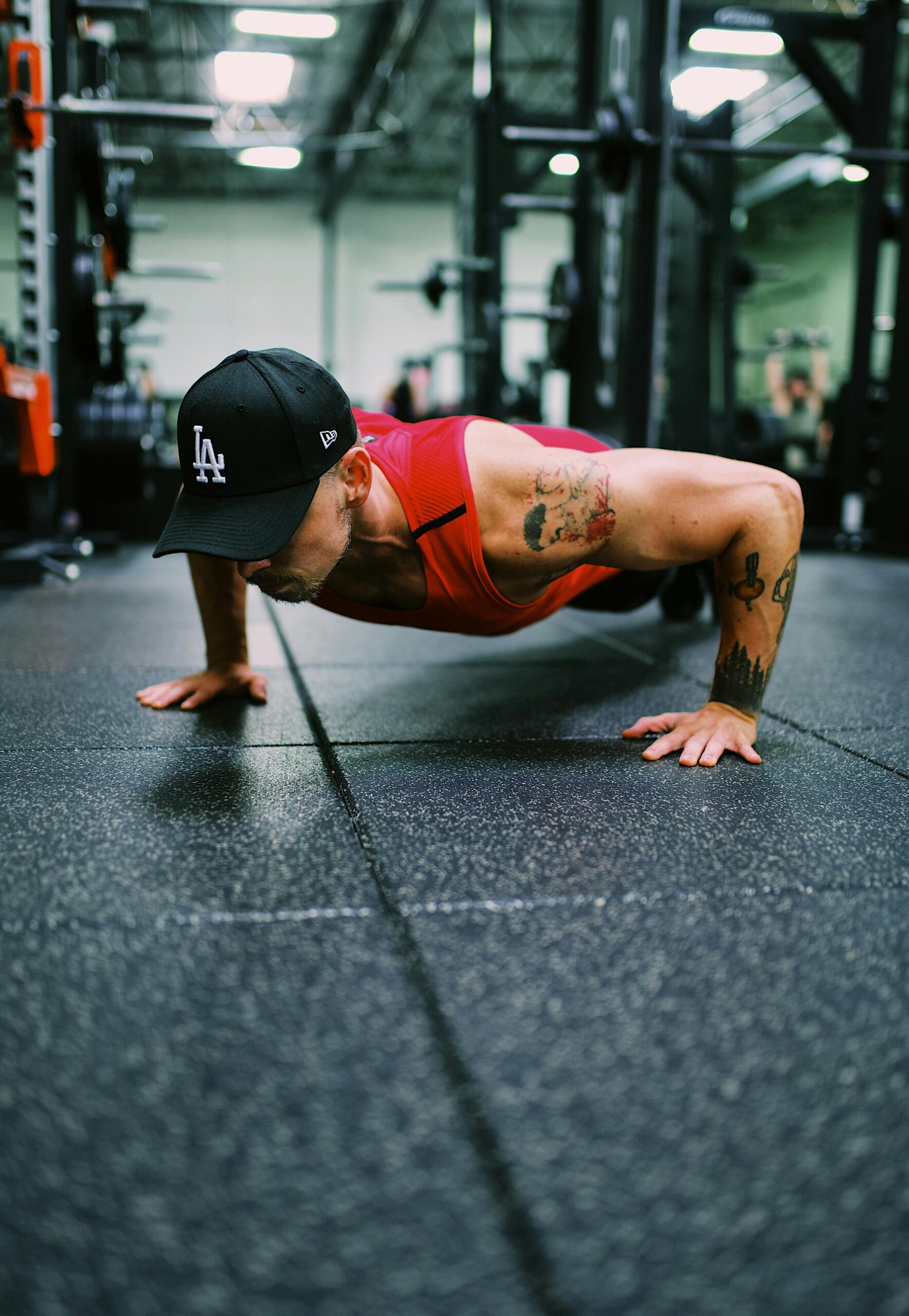 Un hombre tatuado, con una camiseta sin mangas roja y una gorra negra de Los Ángeles, hace una flexión en un gimnasio.