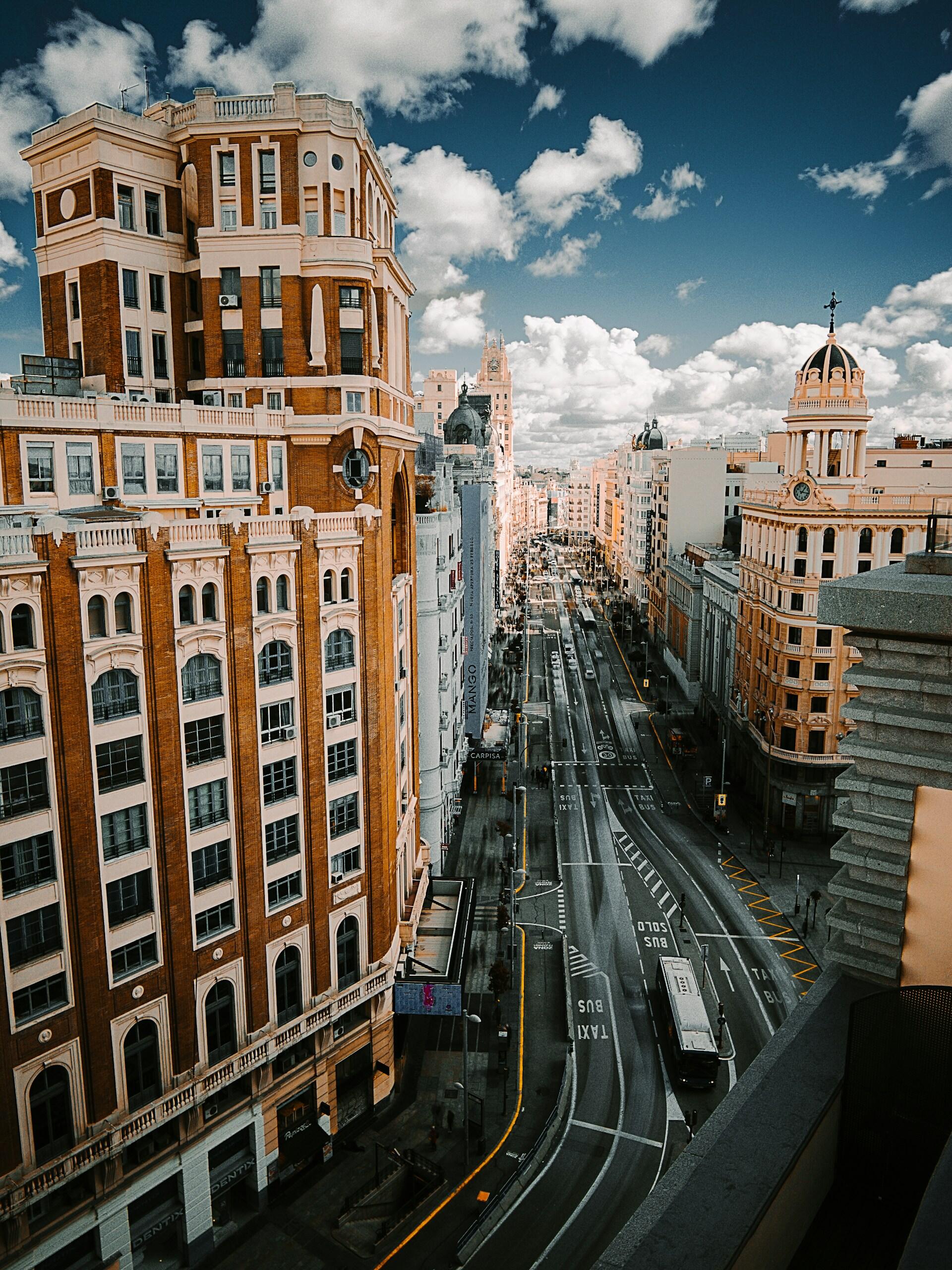 Vista de la Gran Vía de Madrid, con edificios históricos ornamentados, cúpulas, tráfico y unas nubes espectaculares.