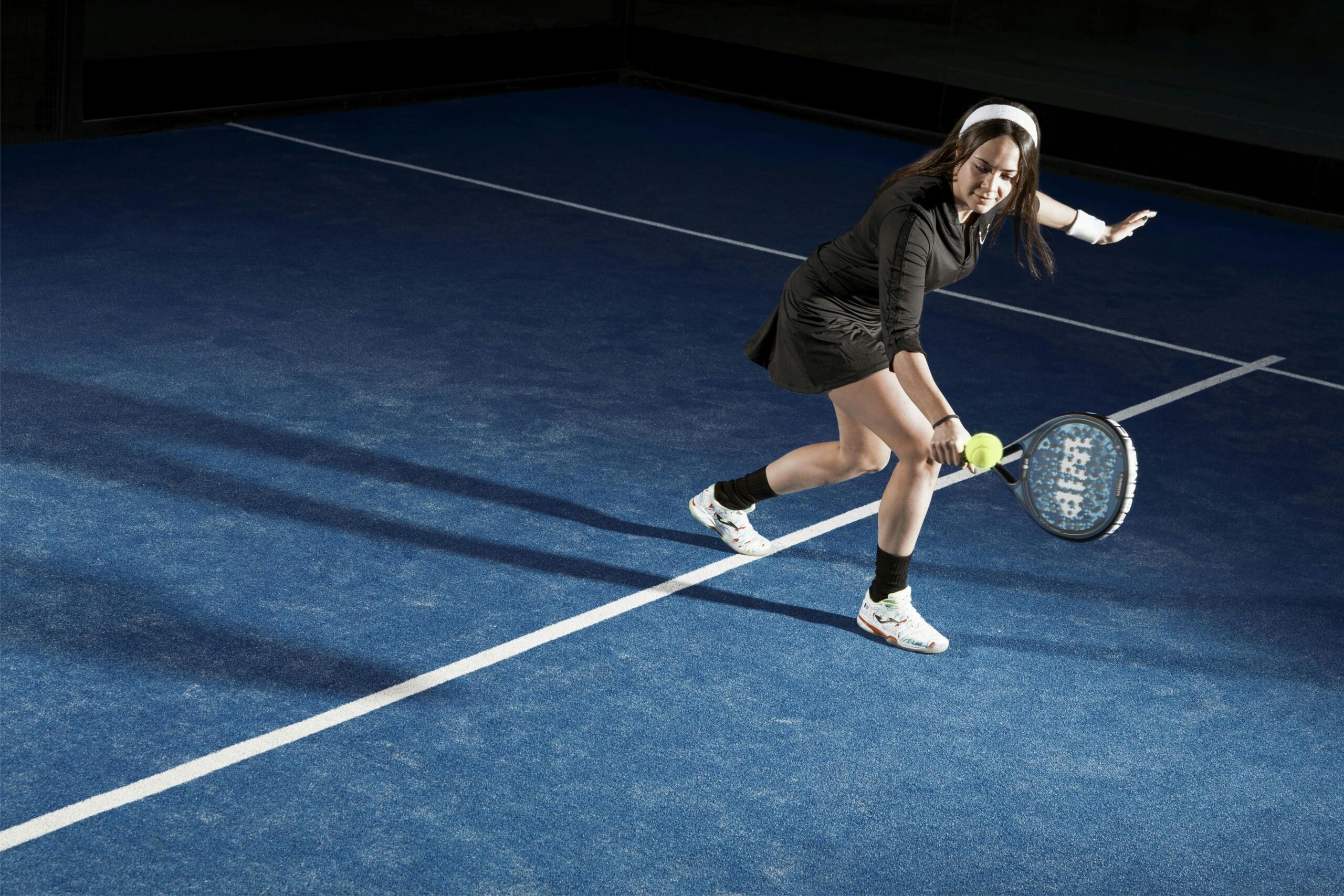 Una mujer vestida con un chándal negro y una cinta blanca en la cabeza golpea una pelota amarilla con una raqueta de pádel en una pista azul con líneas de delimitación blancas, por la noche.