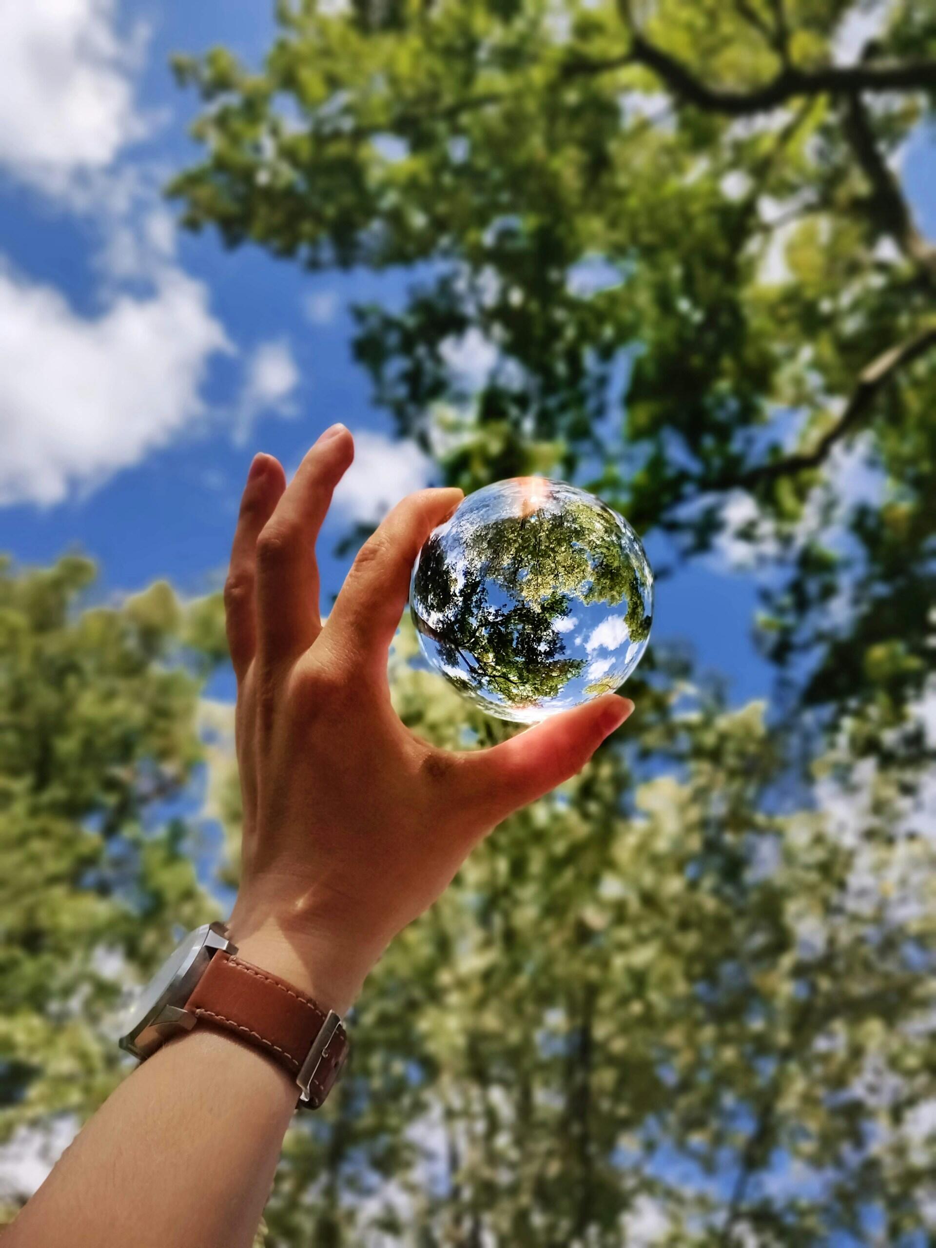Una mano, que lleva un reloj de cuero marrón, sostiene una esfera de cristal frente a un cielo azul, en el que se reflejan árboles verdes y nubes blancas, en una escena surrealista.