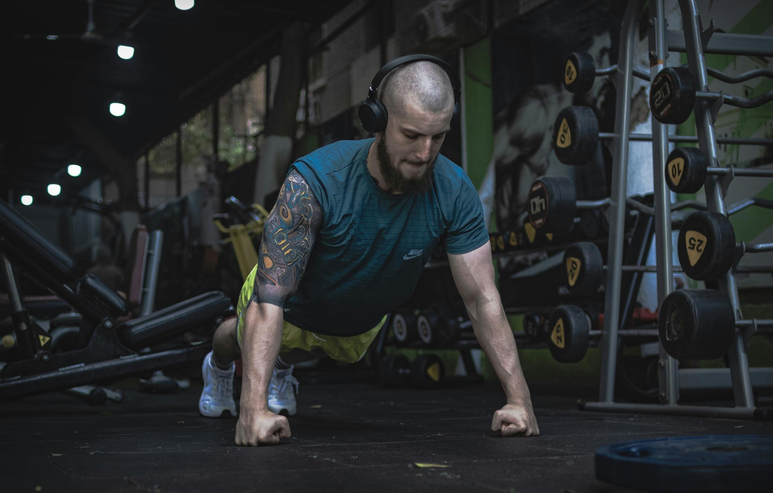 Un hombre con barba y camiseta azul hace flexiones en el suelo de un gimnasio, con soportes para pesas y máquinas de entrenamiento al fondo.
