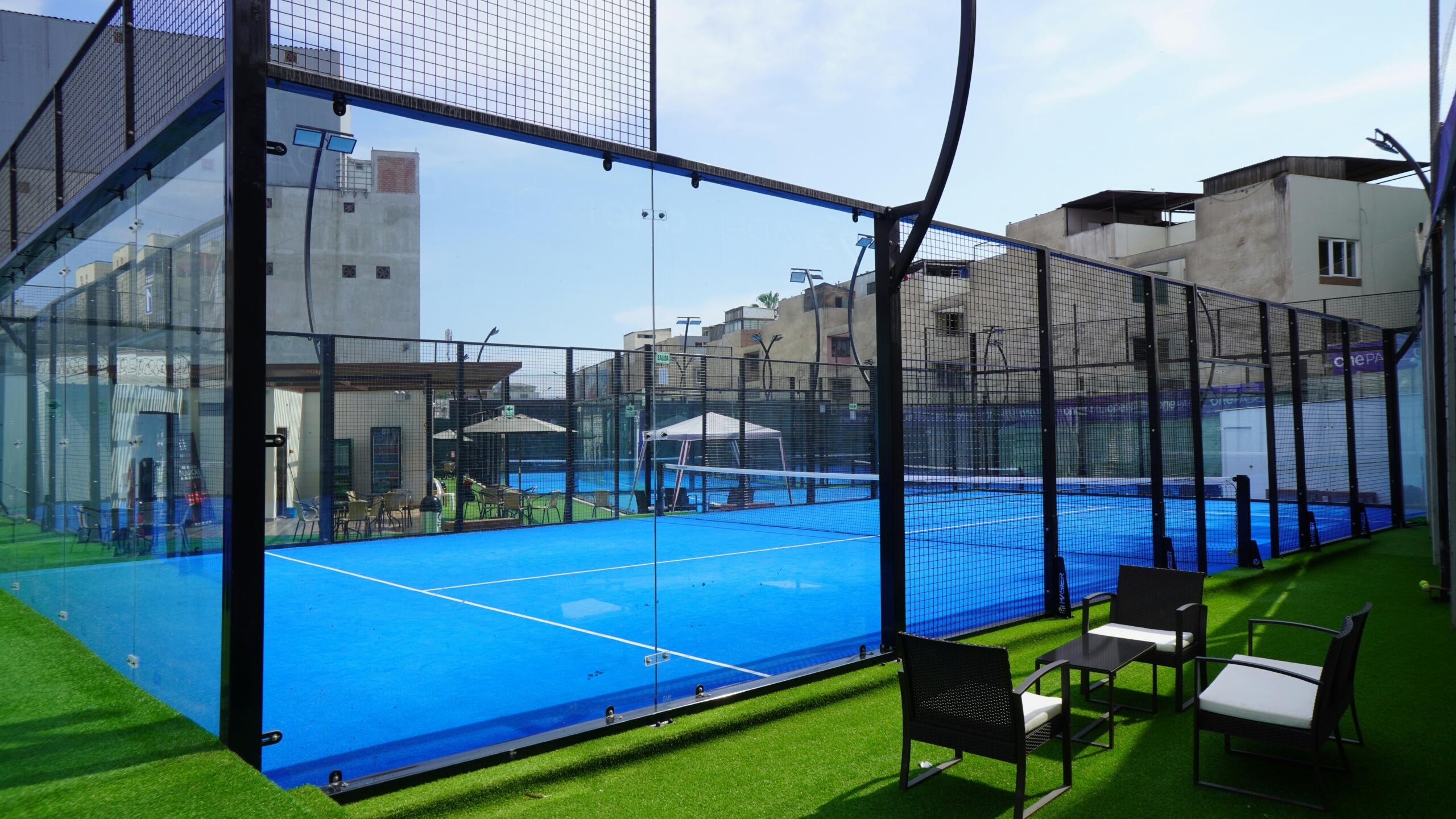 Pista de pádel al aire libre con superficie de juego azul, valla metálica, edificios a su alrededor y tumbonas negras sobre césped artificial.