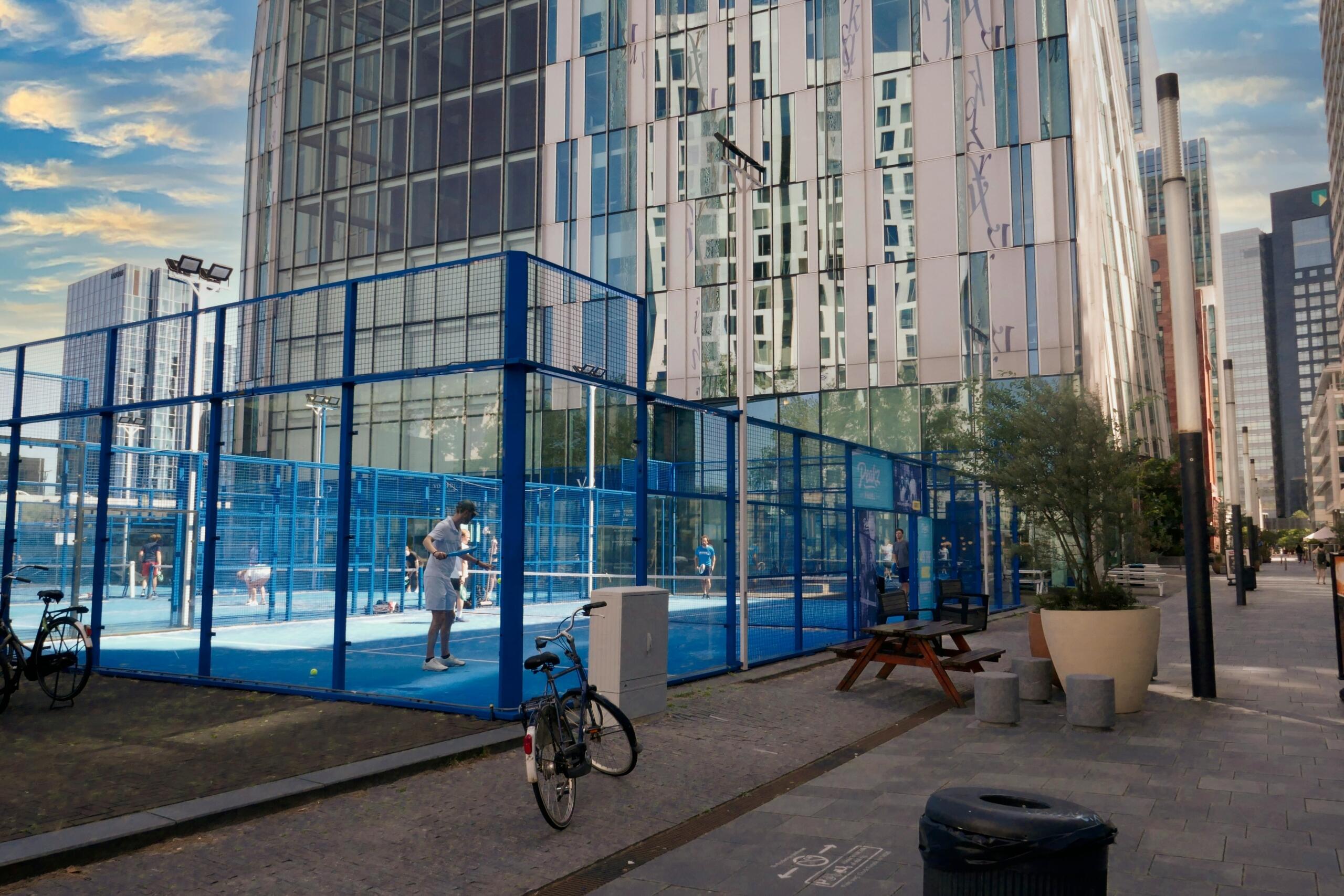 Pista de pádel azul al aire libre rodeada por una valla de malla, con jugadores, bicicletas, bancos y modernos edificios acristalados al fondo.