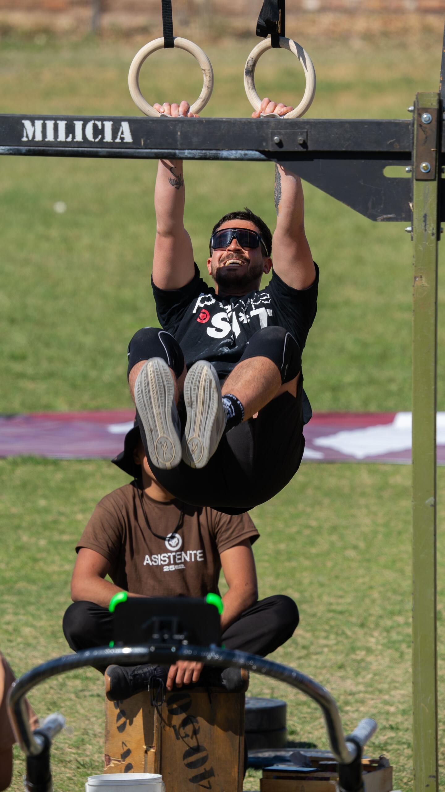 Un deportista sonriente con gafas de sol cuelga de unas anillas de gimnasia de madera en una pista de obstáculos al aire libre.