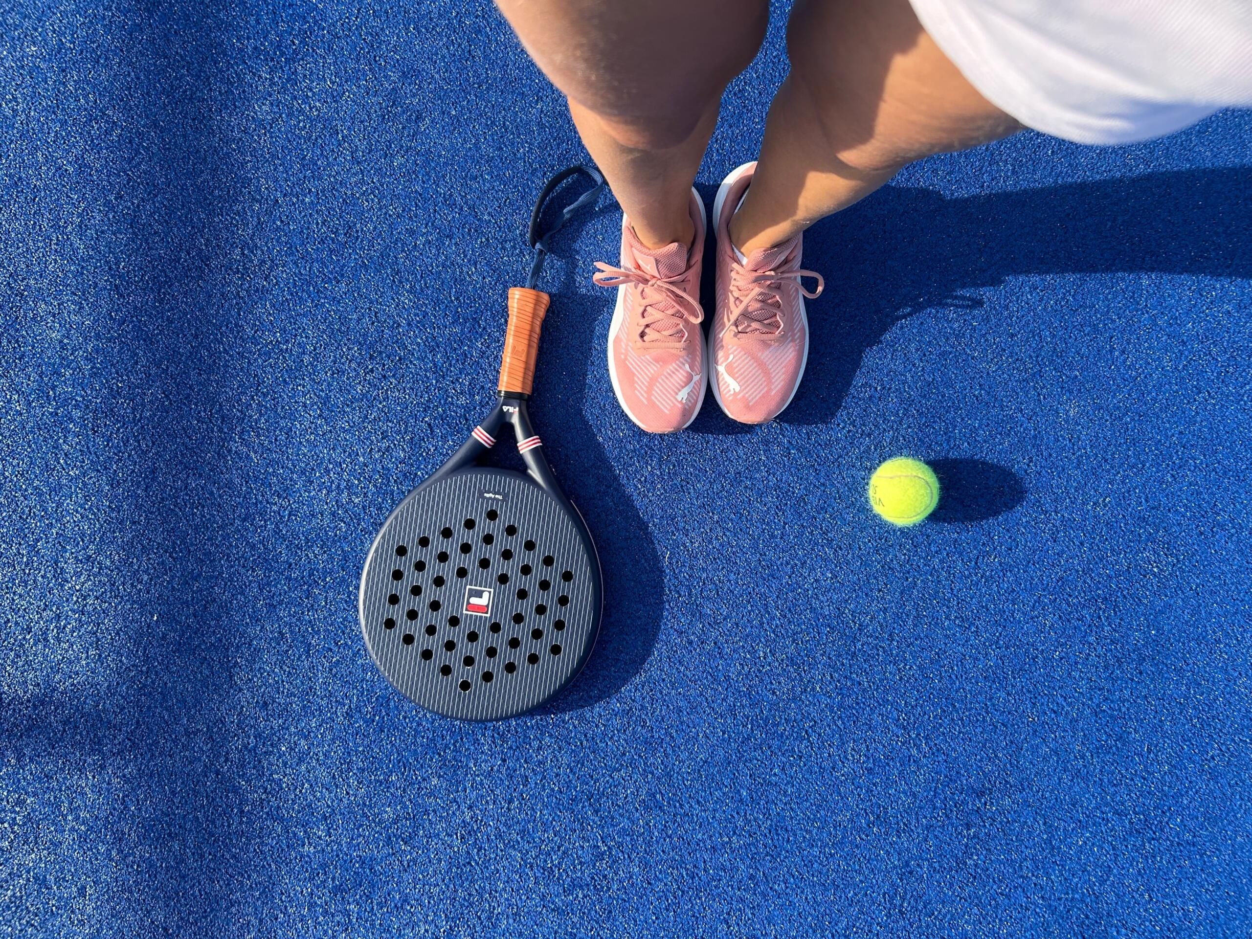 Vista desde arriba de unas zapatillas deportivas rosas, una raqueta de pádel y una pelota de tenis sobre una pista azul.