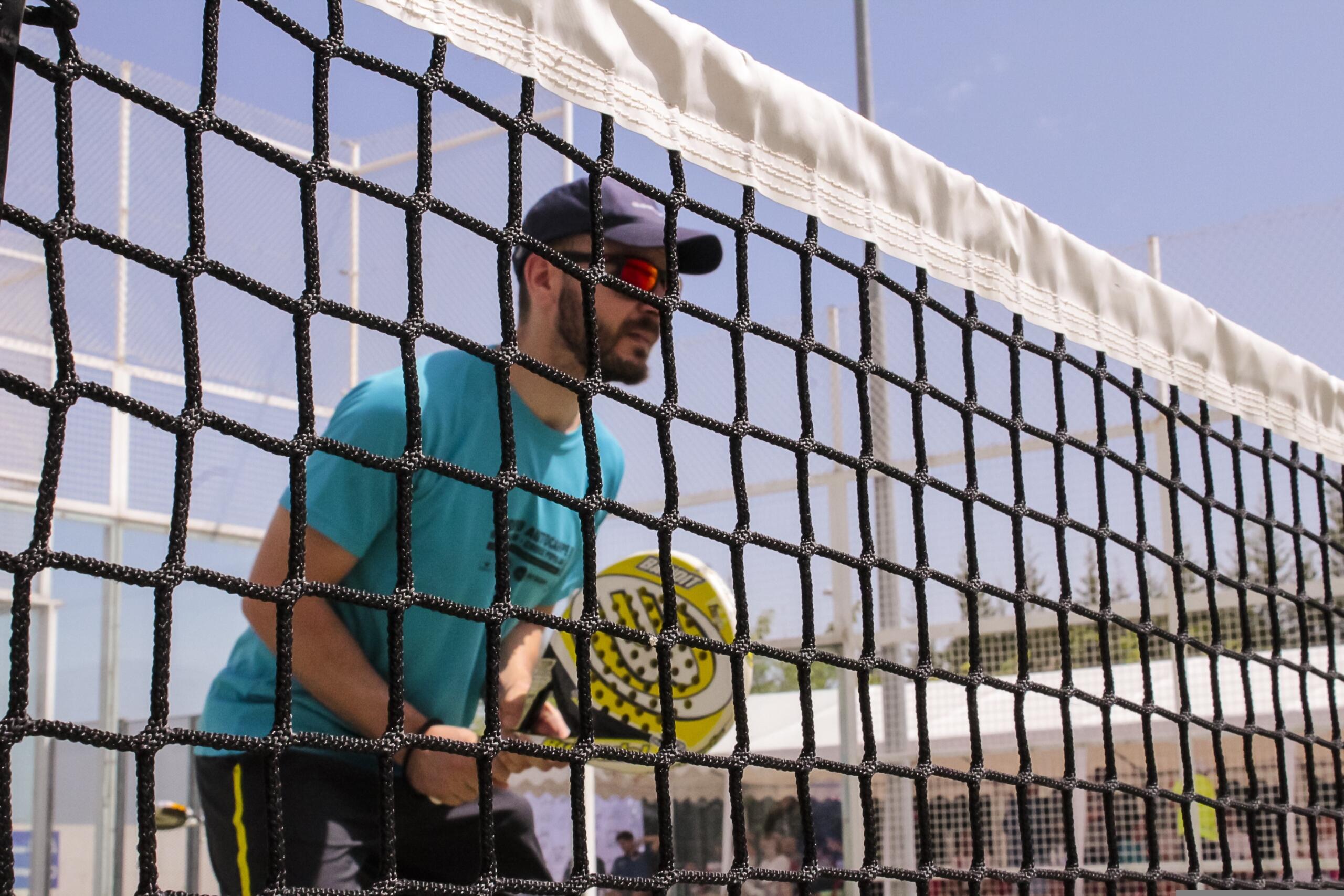 Un hombre con gafas de sol y una gorra está de pie detrás de una red de voleibol negra, bajo una banda blanca de antena.