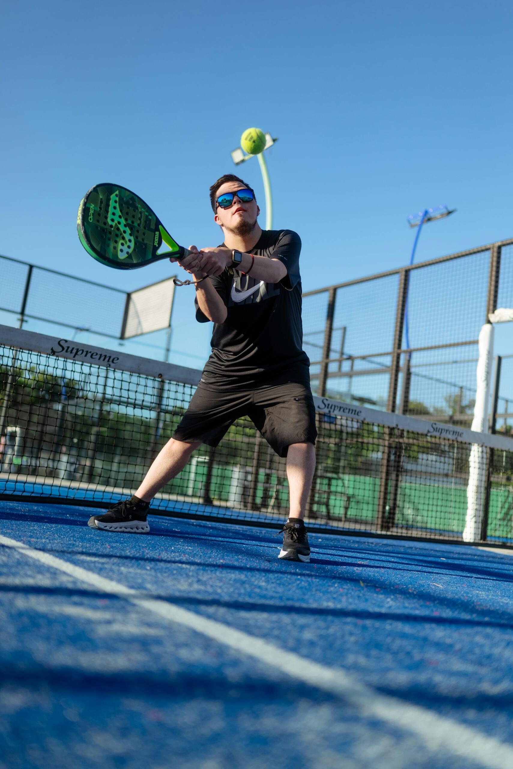 Un hombre con gafas de sol golpea una pelota con una pala verde en una pista de pádel al aire libre.