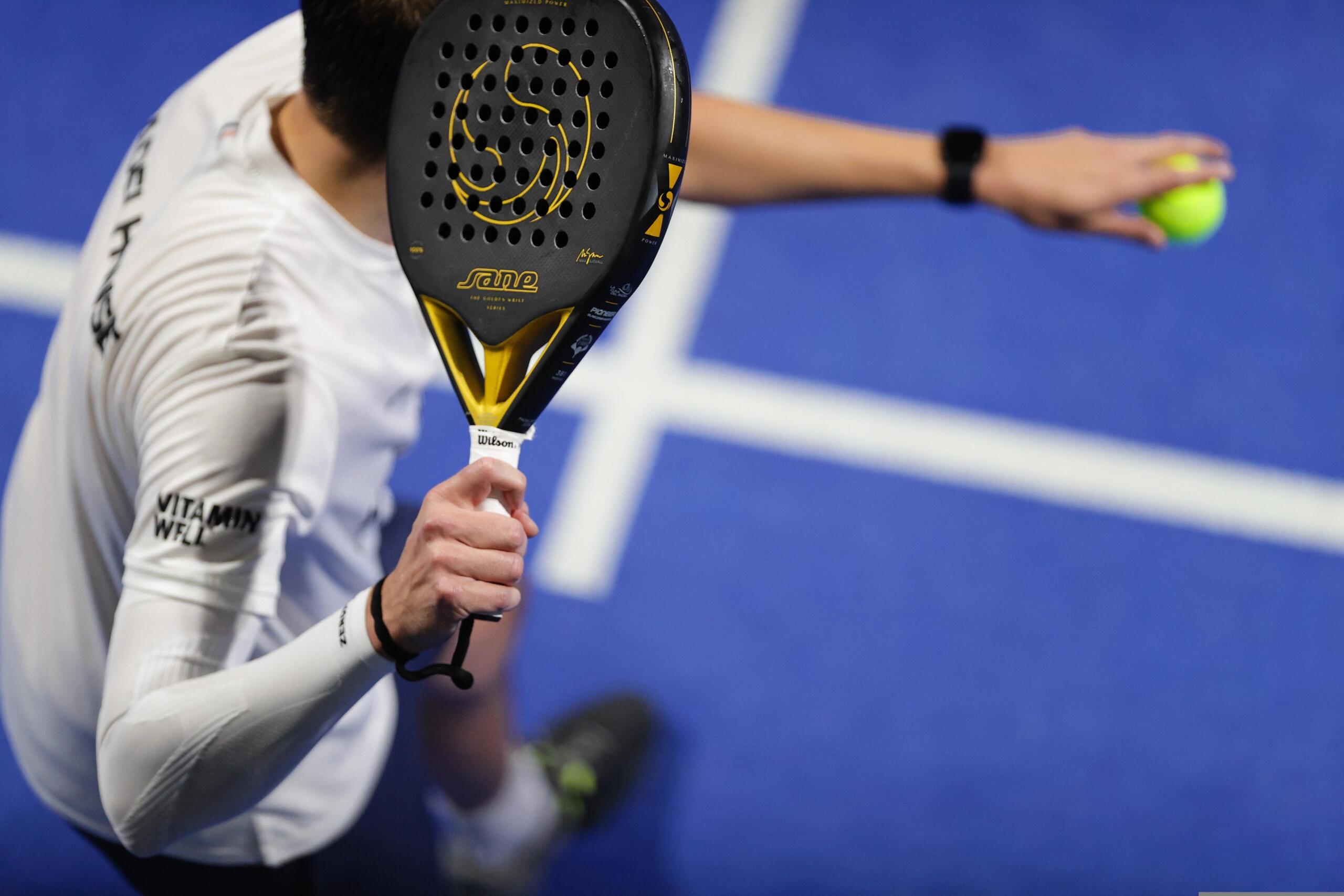 Primer plano de una raqueta deportiva perforada de color negro y amarillo que sostiene una persona con una camiseta blanca.