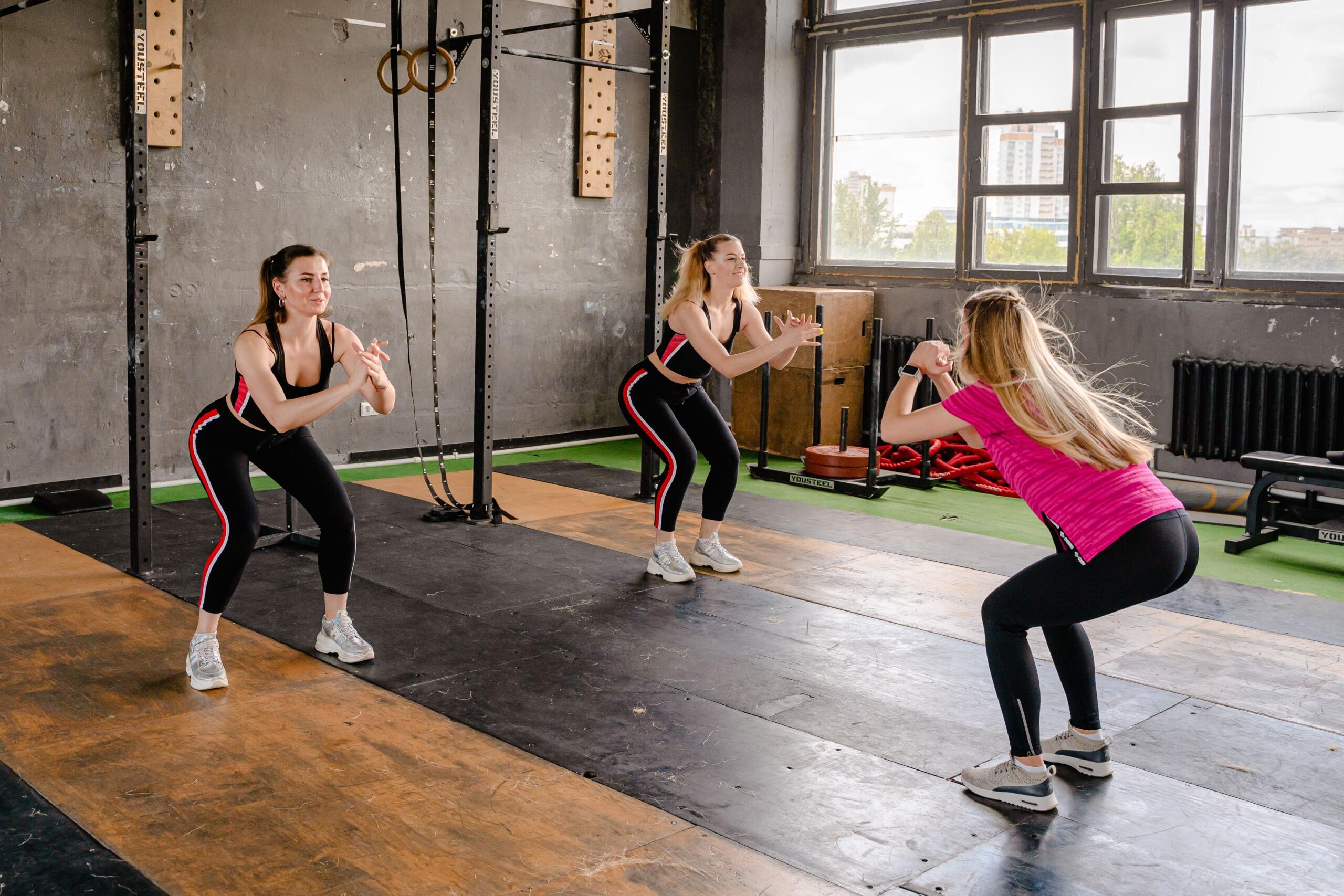 Tres mujeres hacen ejercicio en un gimnasio, realizando sentadillas sincronizadas junto a una jaula de sentadillas y a las máquinas del gimnasio.