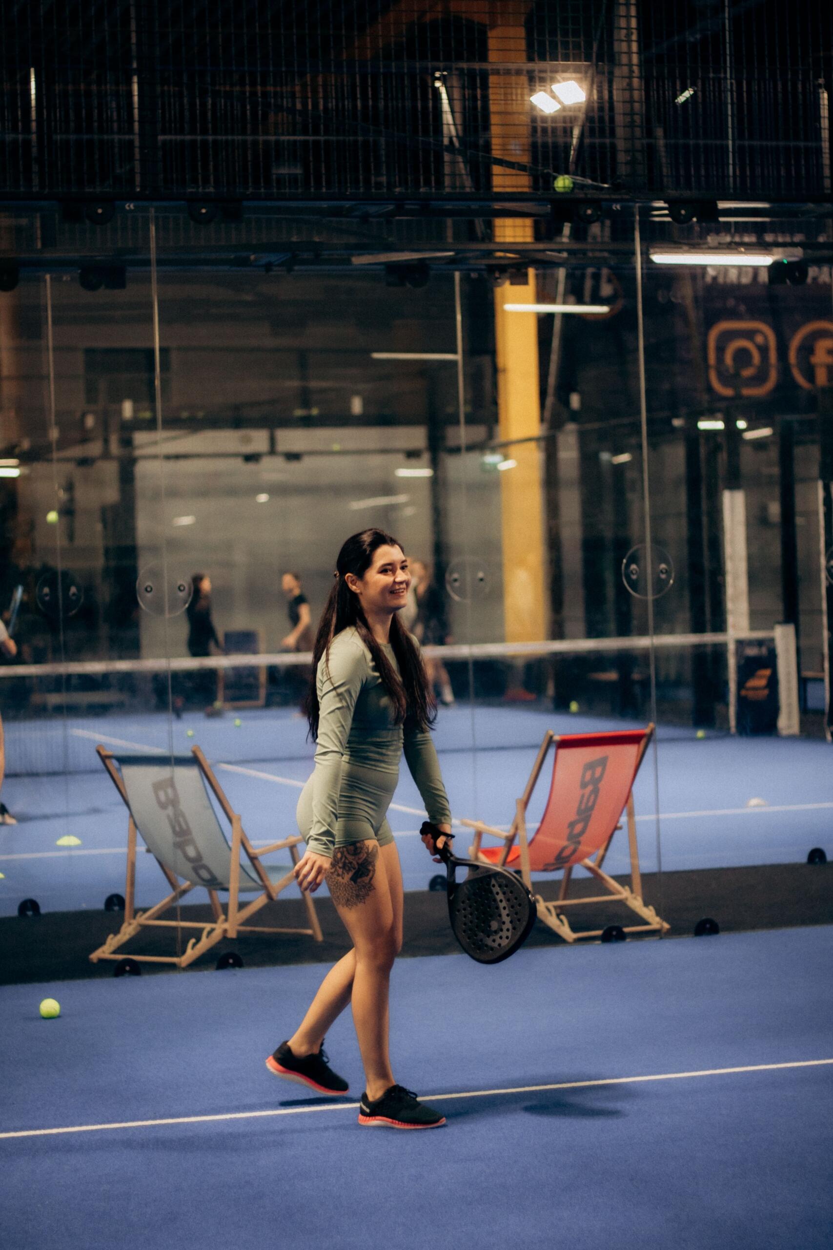 Una mujer vestida con ropa deportiva camina por una pista cubierta de pádel, con una raqueta en la mano y pelotas y tumbonas al fondo.