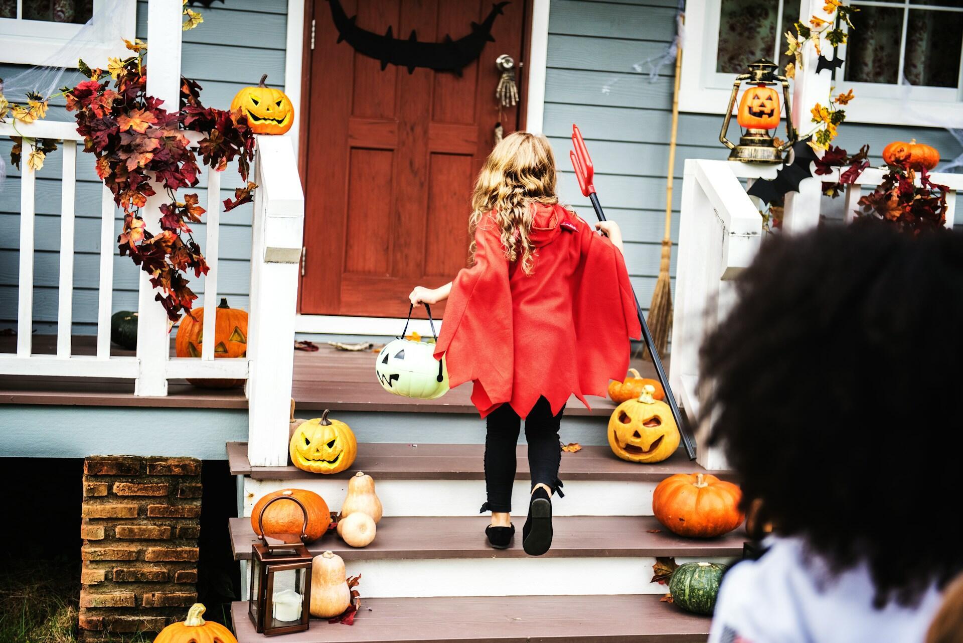 Un niño con un disfraz rojo de Halloween se acerca a un porche decorado con calabazas, llevando una cubeta para pedir caramelos y una horca.