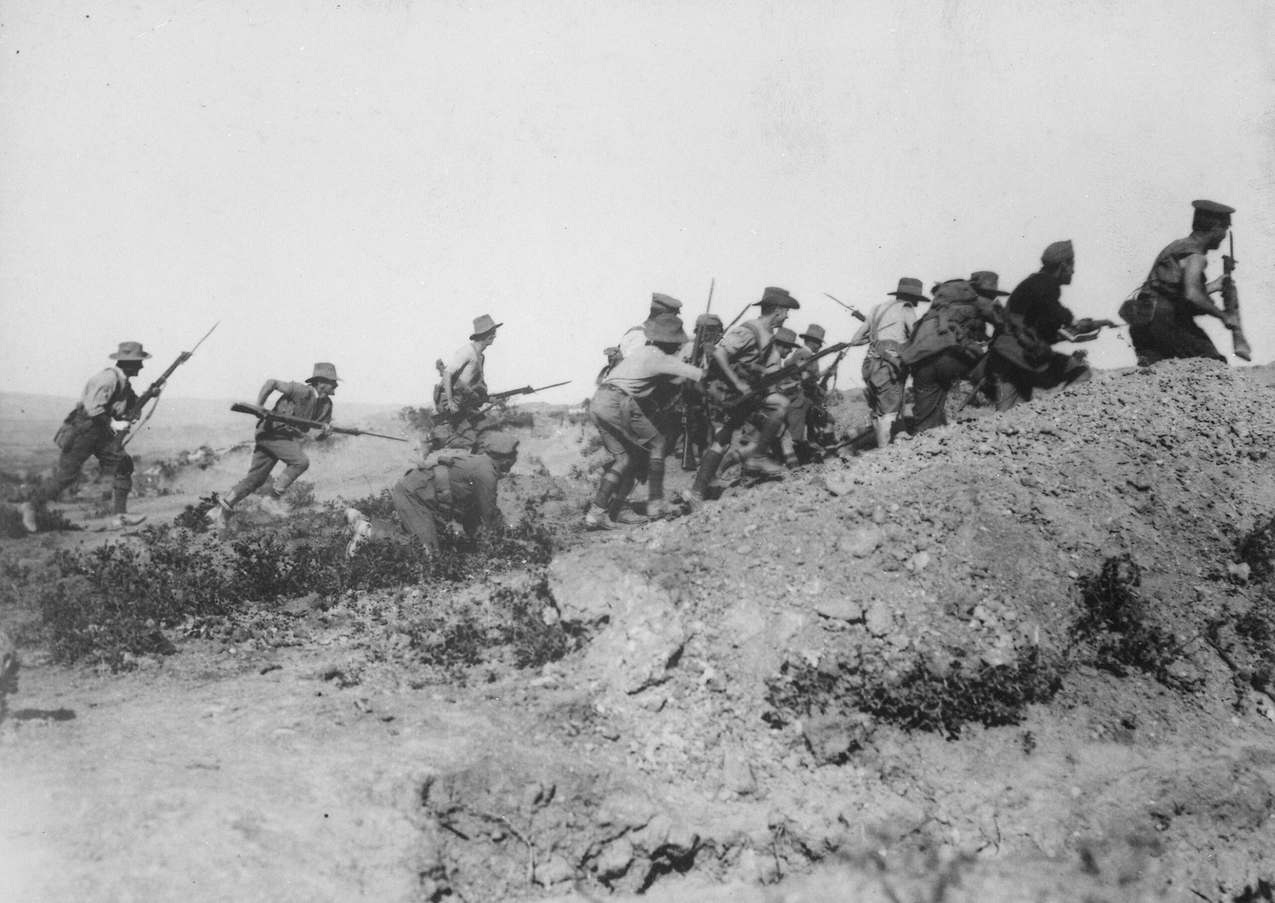 Black-and-white photo of soldiers carrying rifles as they advance uphill over rugged terrain.