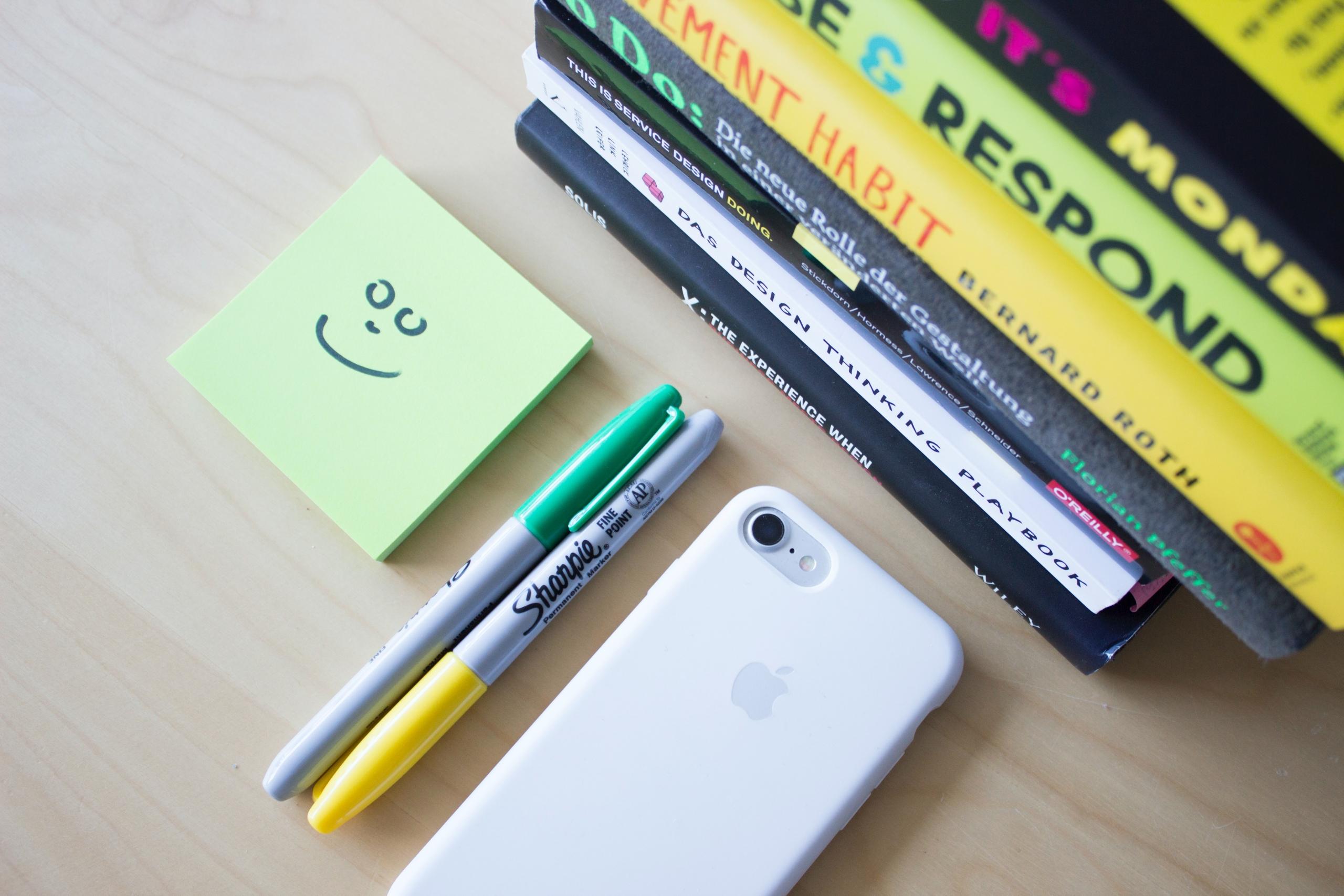 Iphone, post-it-lappuja, kaksi kynää ja kirjoja pöydällä