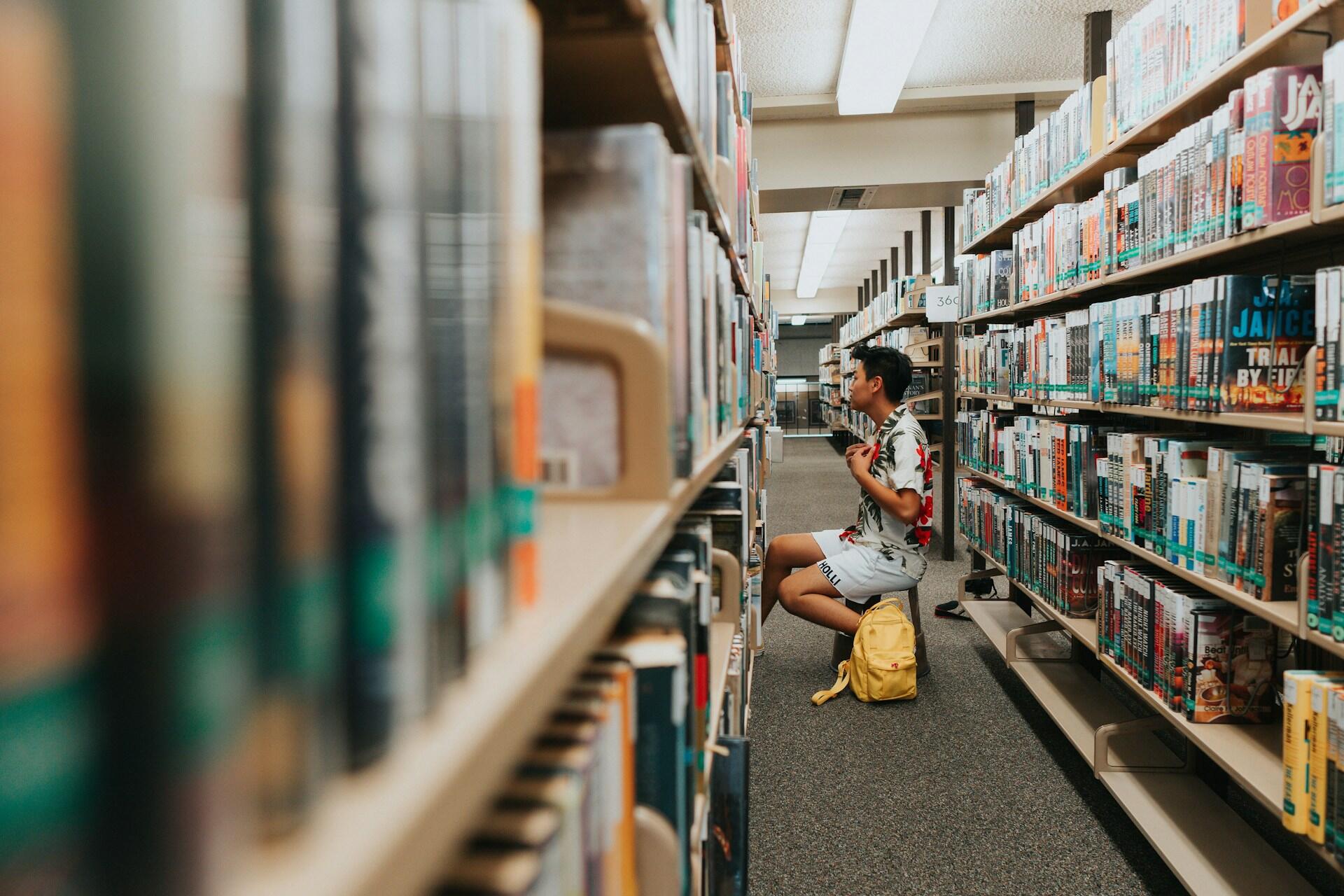 Mies istumassa penkillä kirjahyllyjen välissä