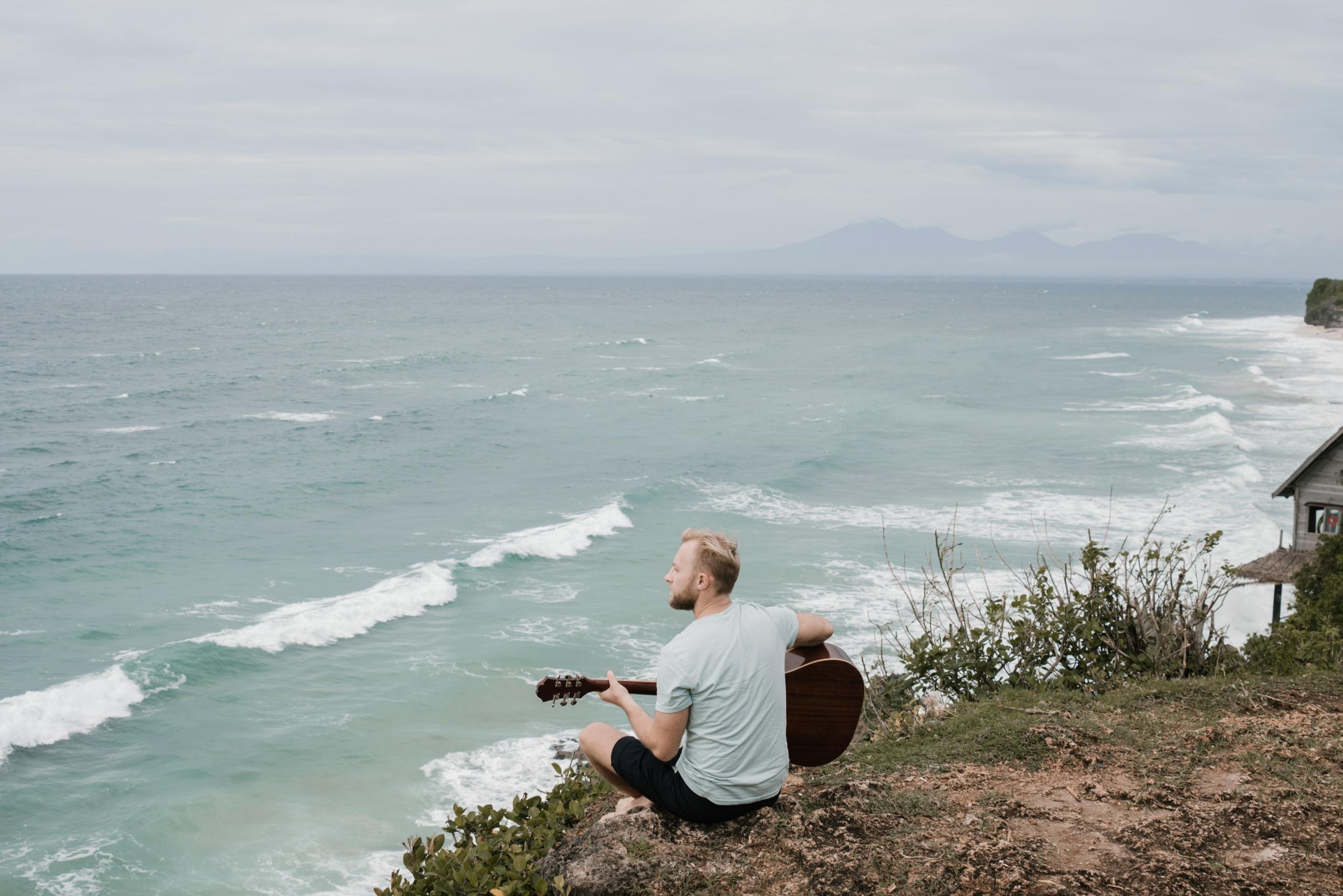 Jeune homme blond de dos une guitare à la main, face à l'océan. 