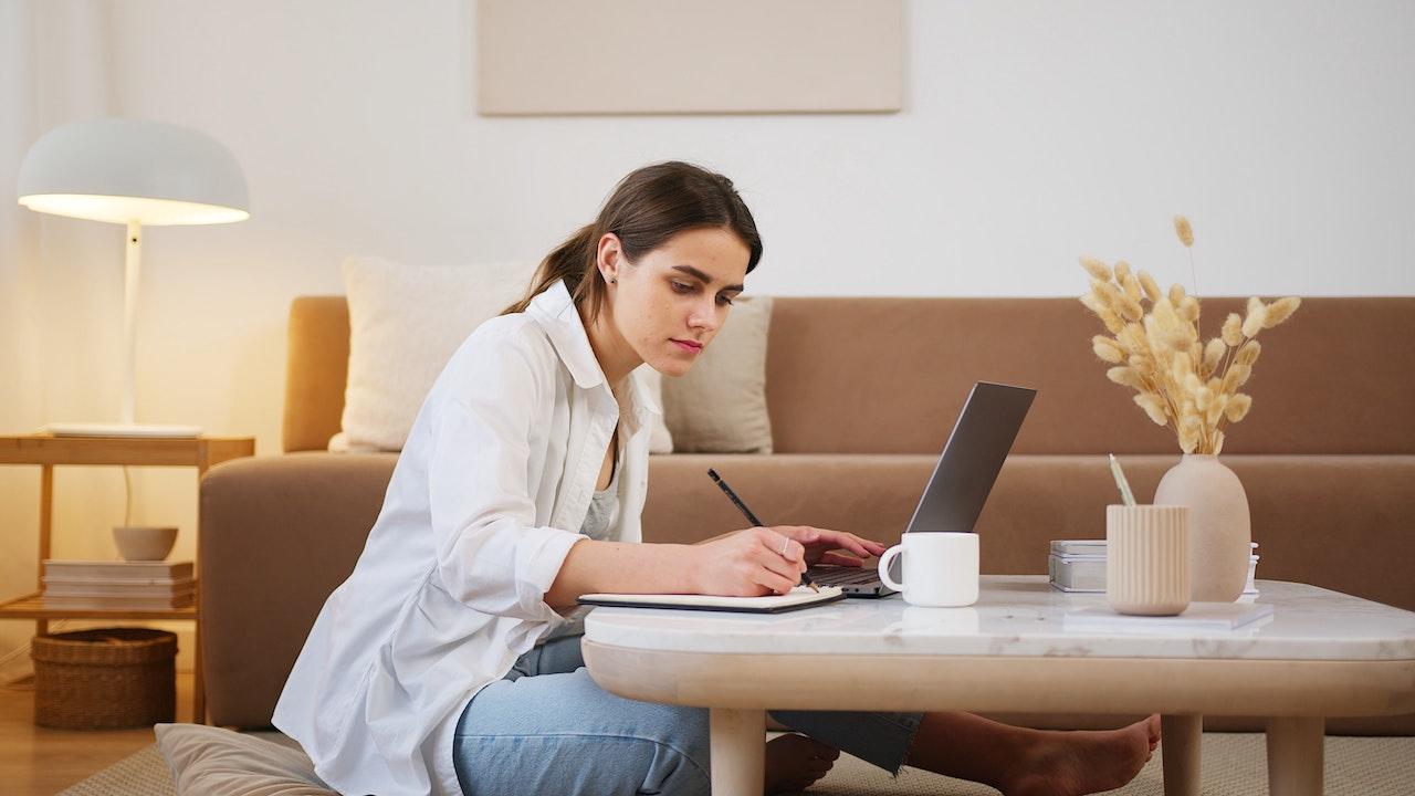 Jeune femme assise par terre : elle écrit sur un carnet sur sa table basse avec un ordinateur ouvert et une tasse de café. 