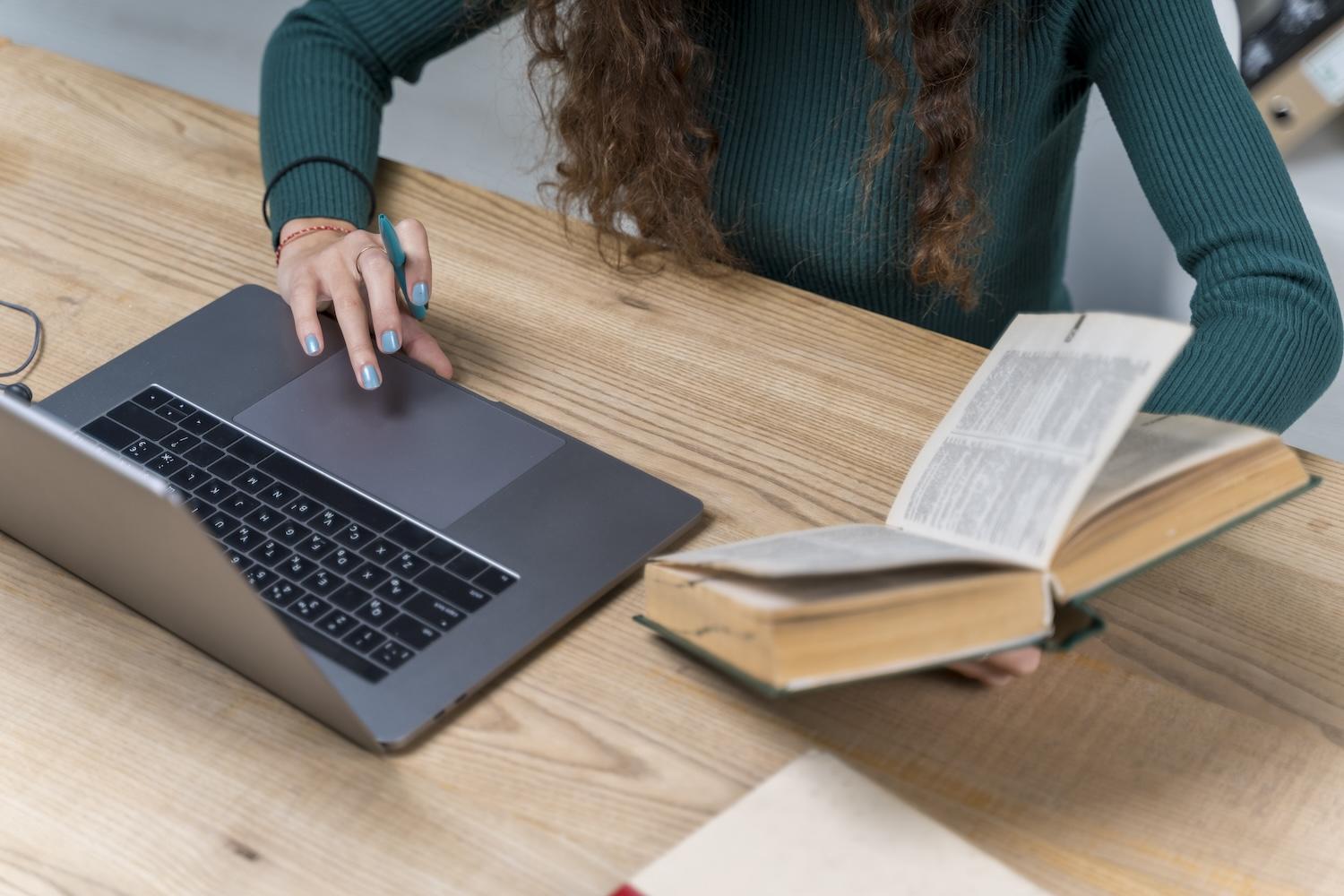 Une jeune femme travaille sur un ordinateur avec un livre dans la main.