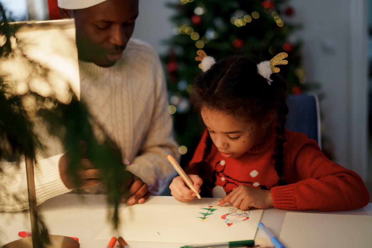 Une petite fille qui dessine un père noël et un sapin aux côtés de son père. 