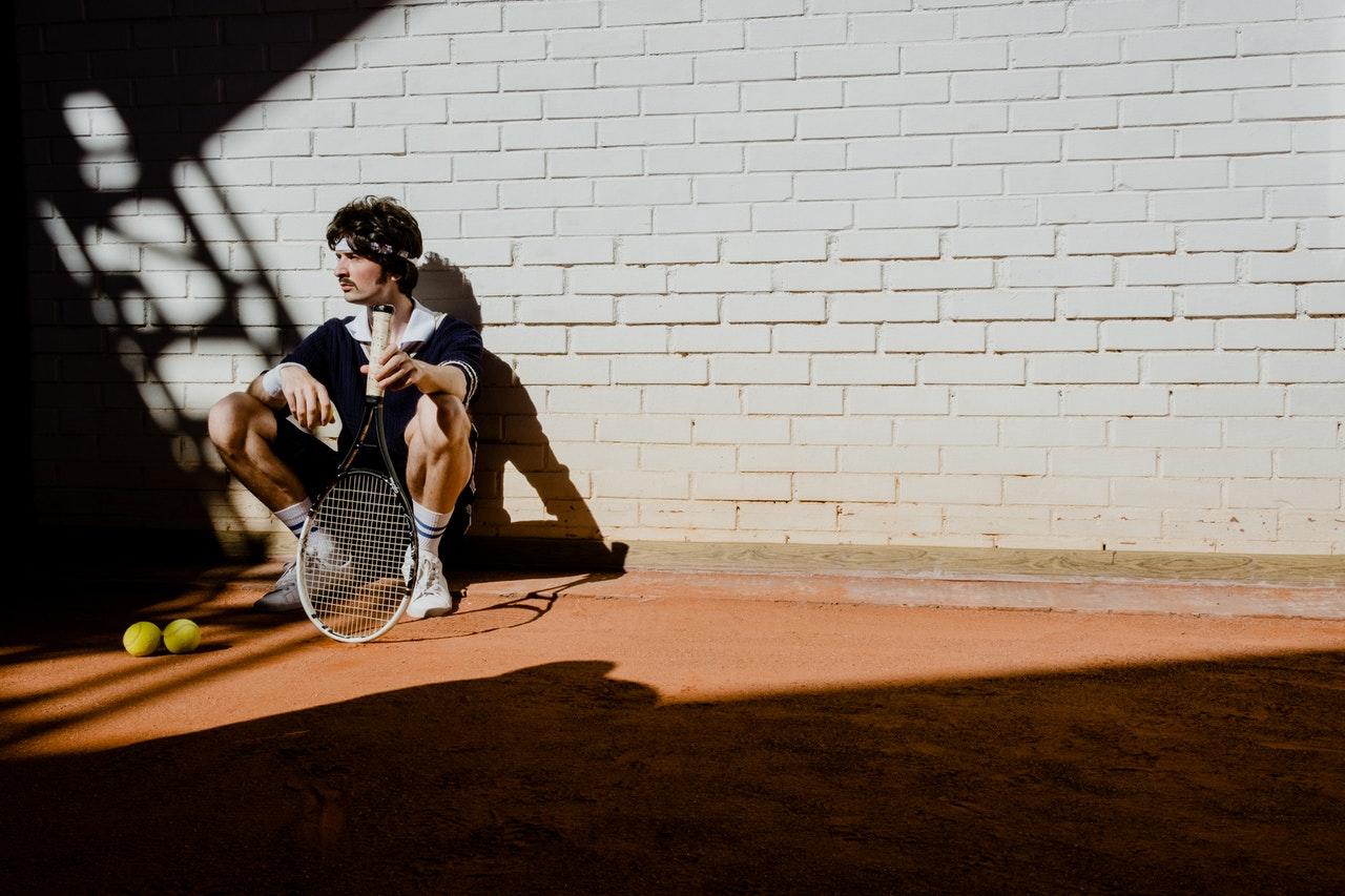 Jeune homme accroupi contre le mur d'un court de tennis en terre battue, tenant une raquette. 
