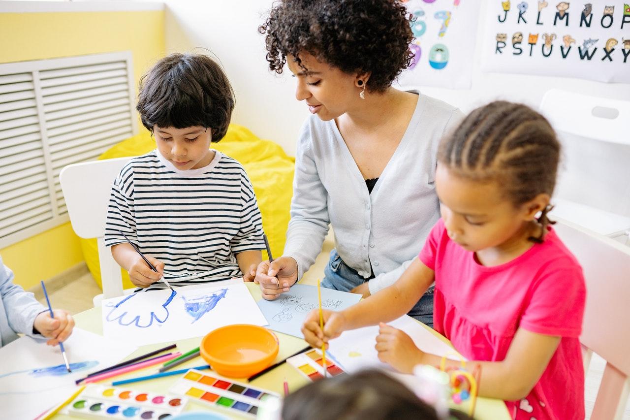 Enseignante d'école maternelle entourée de deux enfants qui dessinent.