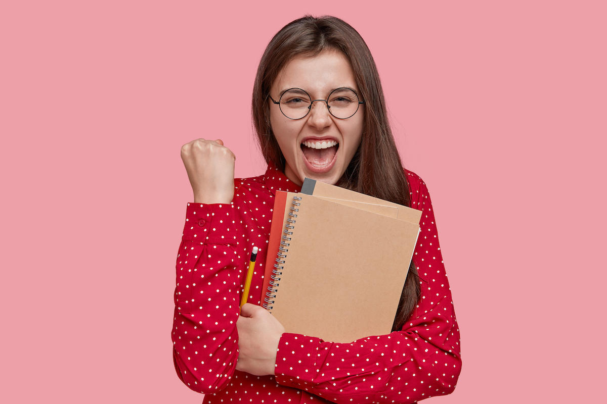 Jeune femme tenant des cahiers et un crayon dans la main le poing fermé et à l'expression heureuse.