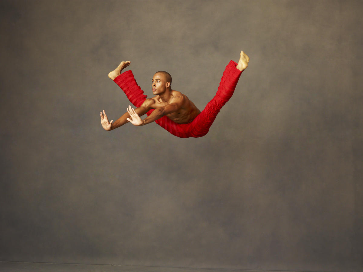 Alvin Ailey en saut jambes écartées, pieds flex, buste vers l'avant et bras tendus au milieu des jambes avec les mains flex.