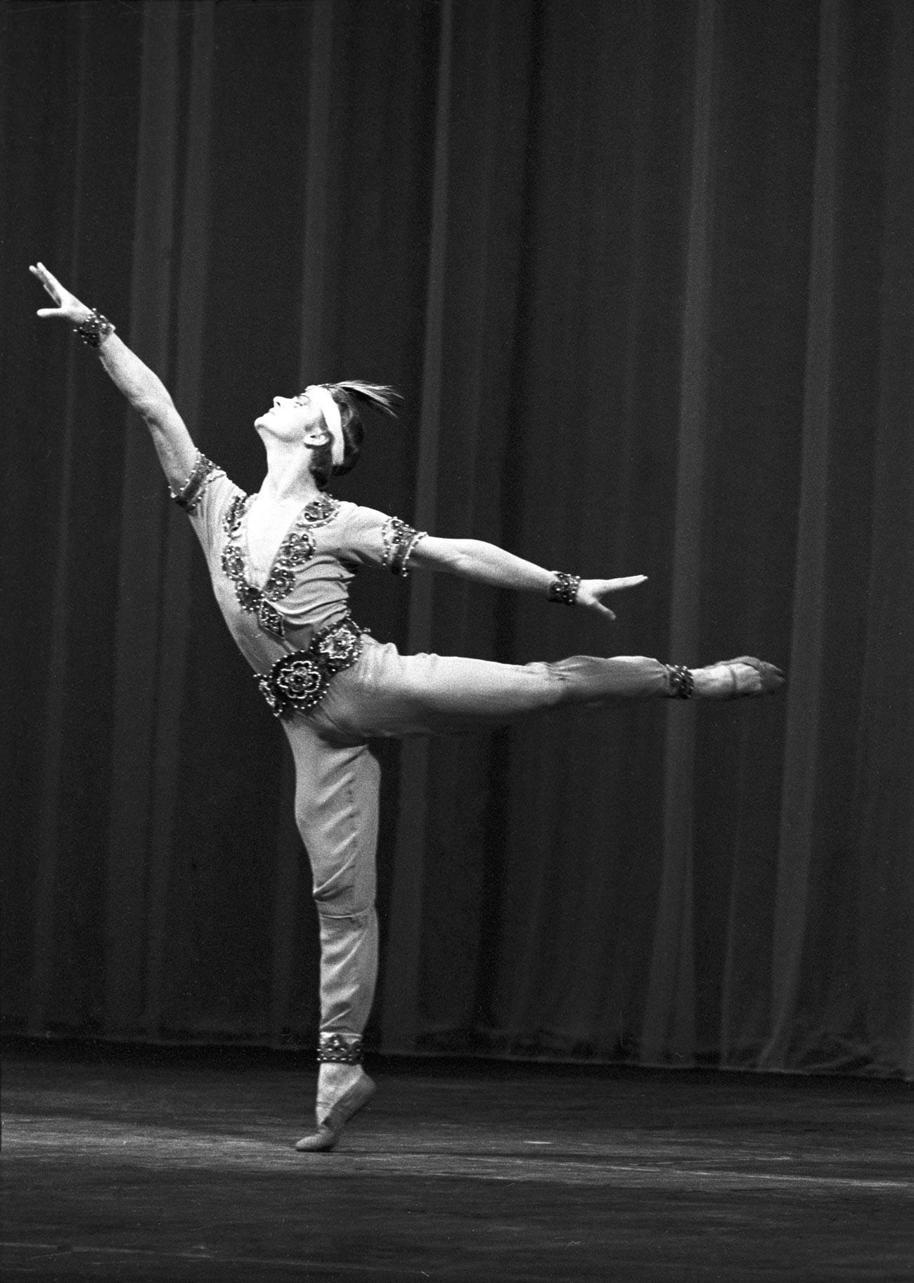 Mikhaïl Baryshnikov sur demi-pointe sur la jambe droite, jambe gauche tendue parallèle au sol en arrière, bras tendus et tête relevée.
