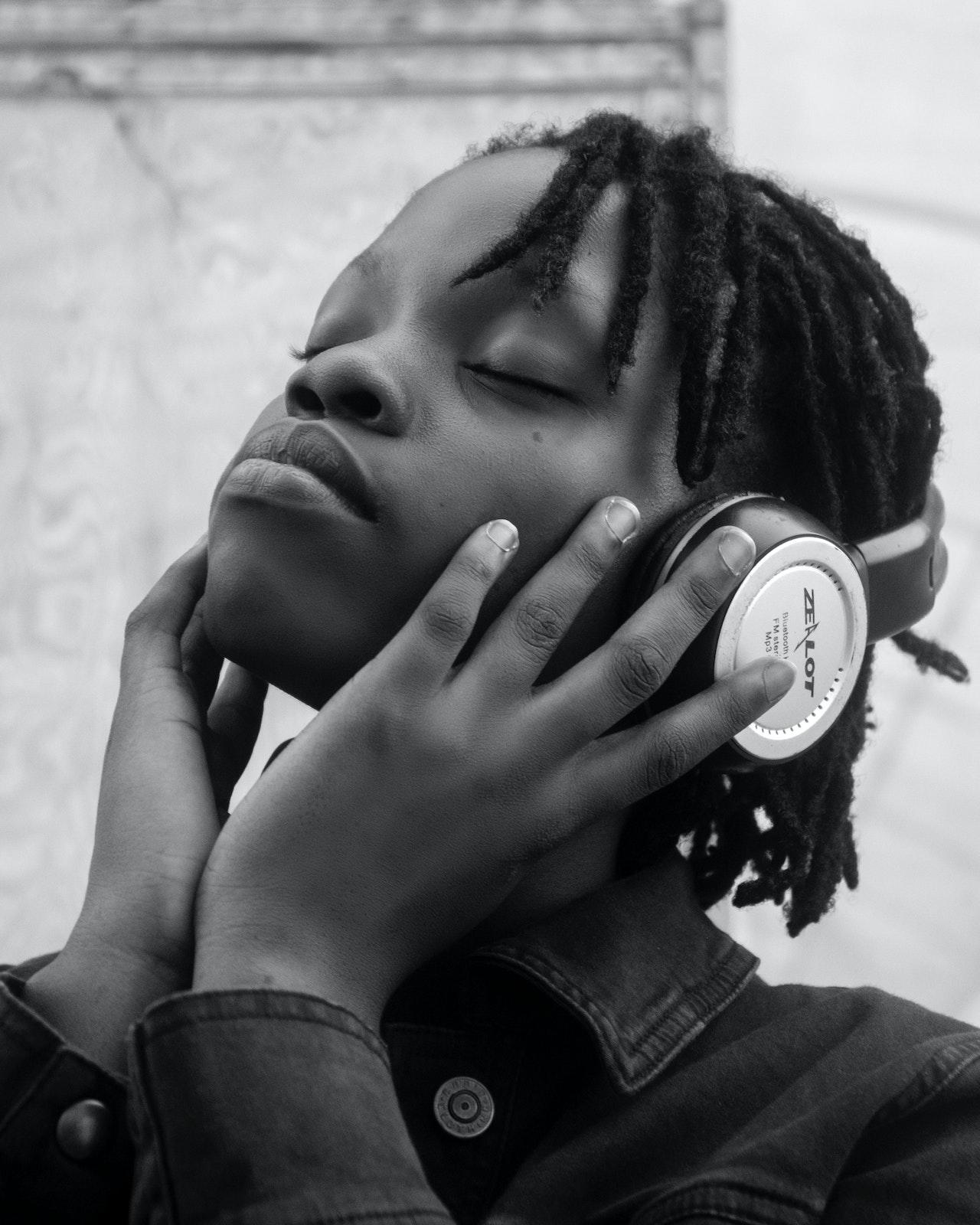 Jeune femme noire avec un casque sur les oreilles en train d'écouter de la musique les yeux fermés et les mains sur son visage. 