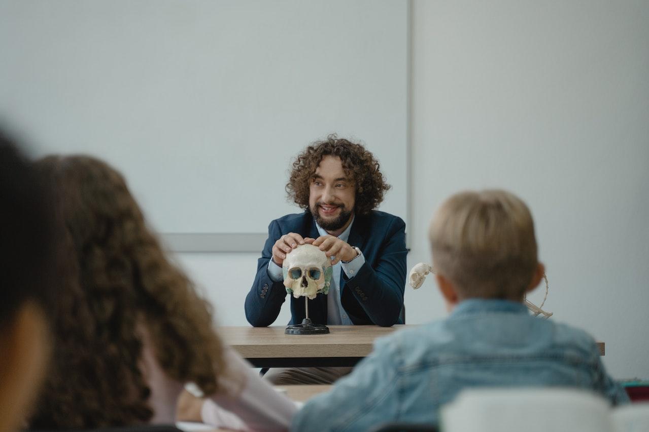 Professeur assis à son bureau avec les mains posées sur un crâne. On voit les élèves de dos.