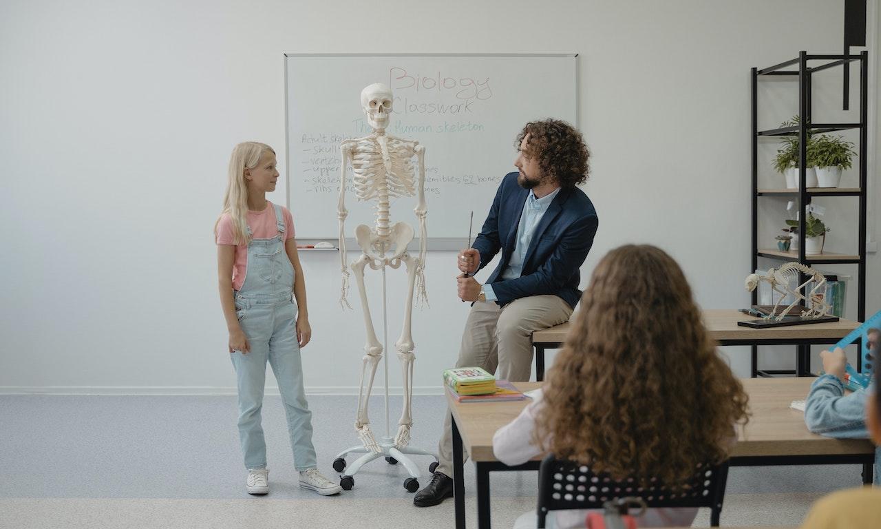 Professeur aux cheveux bouclés interroge une élève blonde devant le squelette d'un être humain dans une salle de classe.