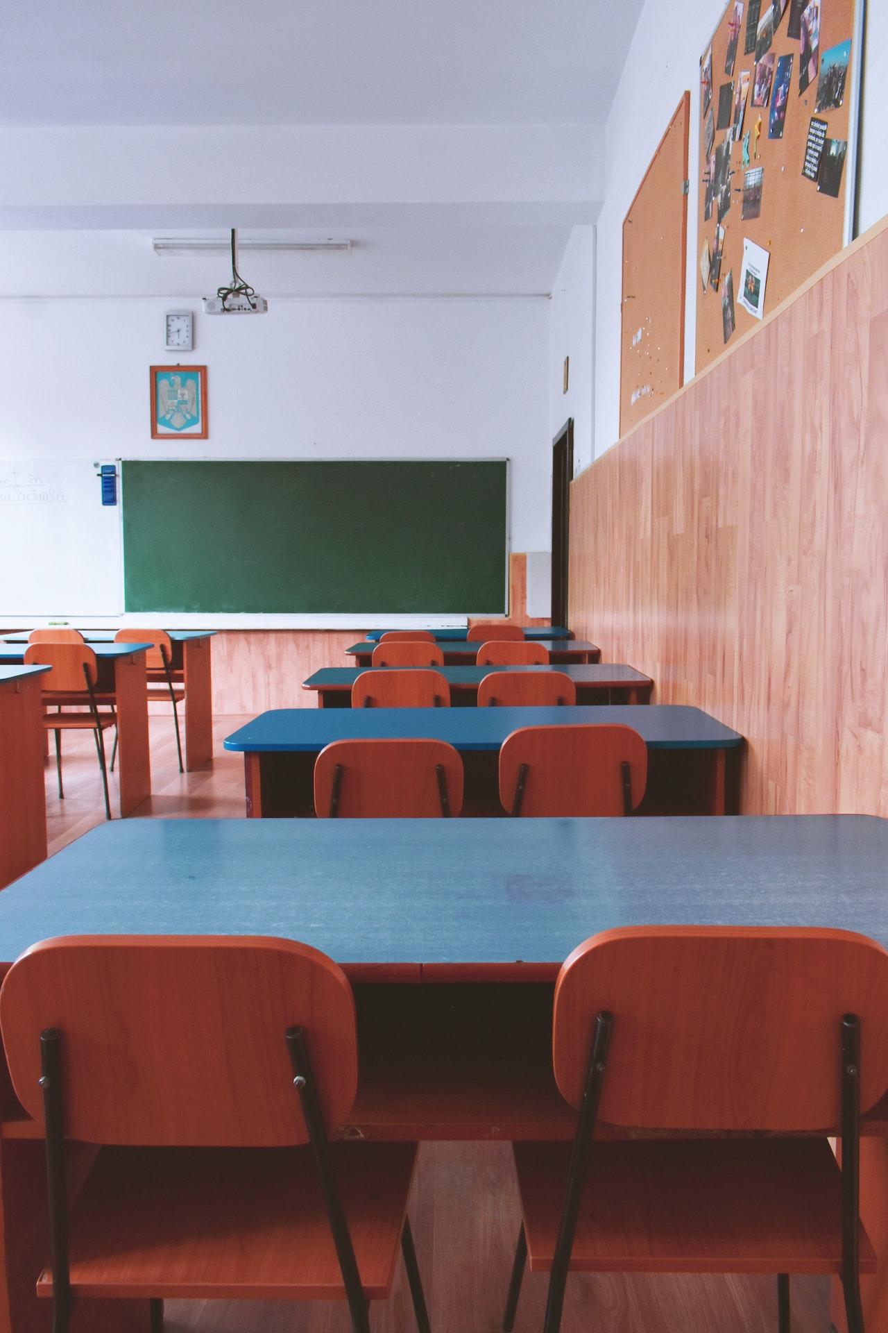 Salle de classe vide avec un tableau vert au fond et des tables et chaises au premier plan.