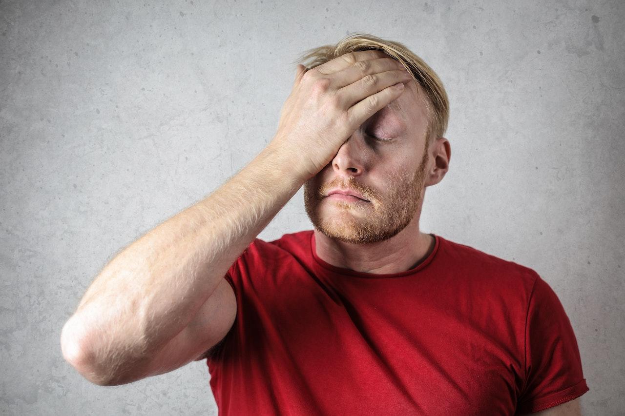 Homme blanc et blond vénitien, d'environ 35-40 ans qui se tient la tête. Il porte un t-shirt rouge. 