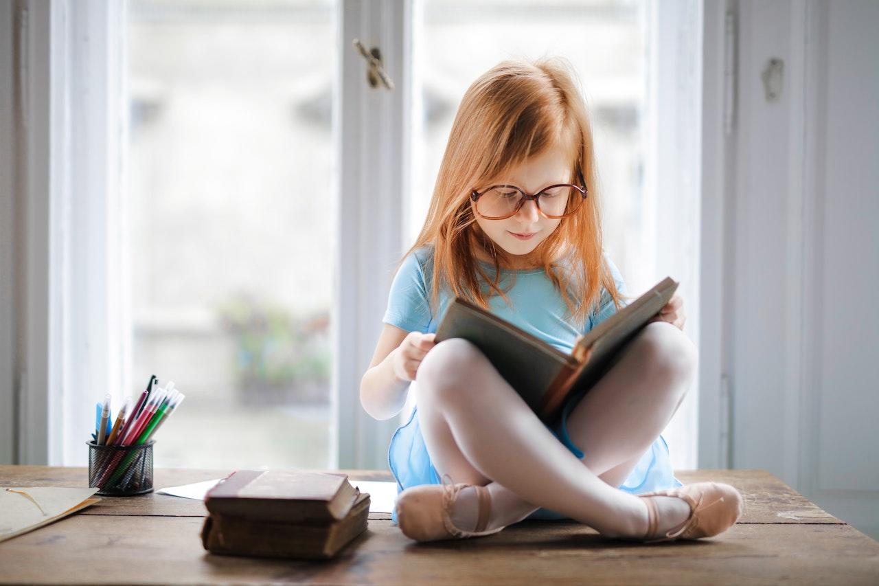 Petite fille rousse assise en tailleur sur son bureau, un livre entre les mains.